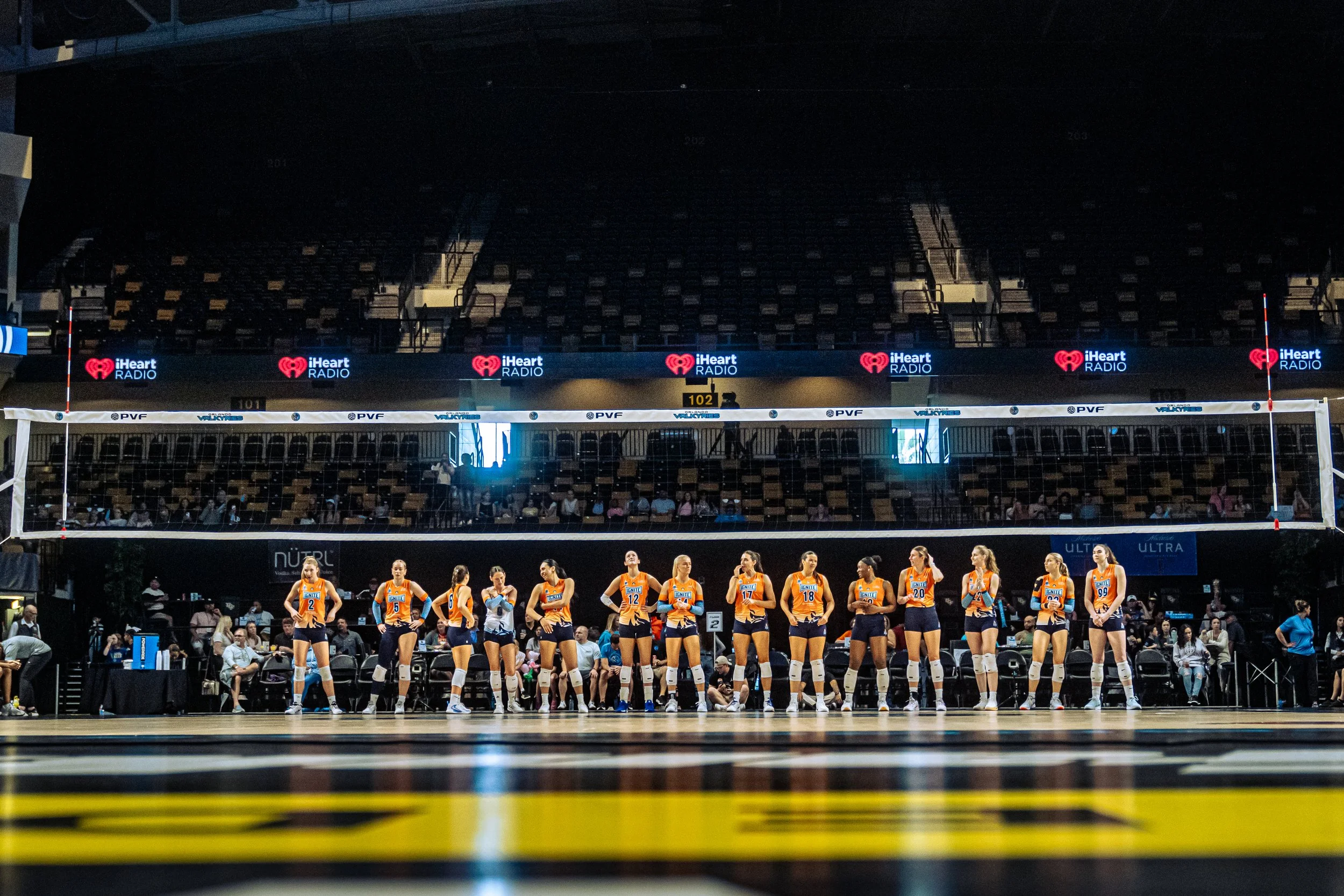 A women's volleyball team standing in a line on an indoor court during a game with spectators in the background.