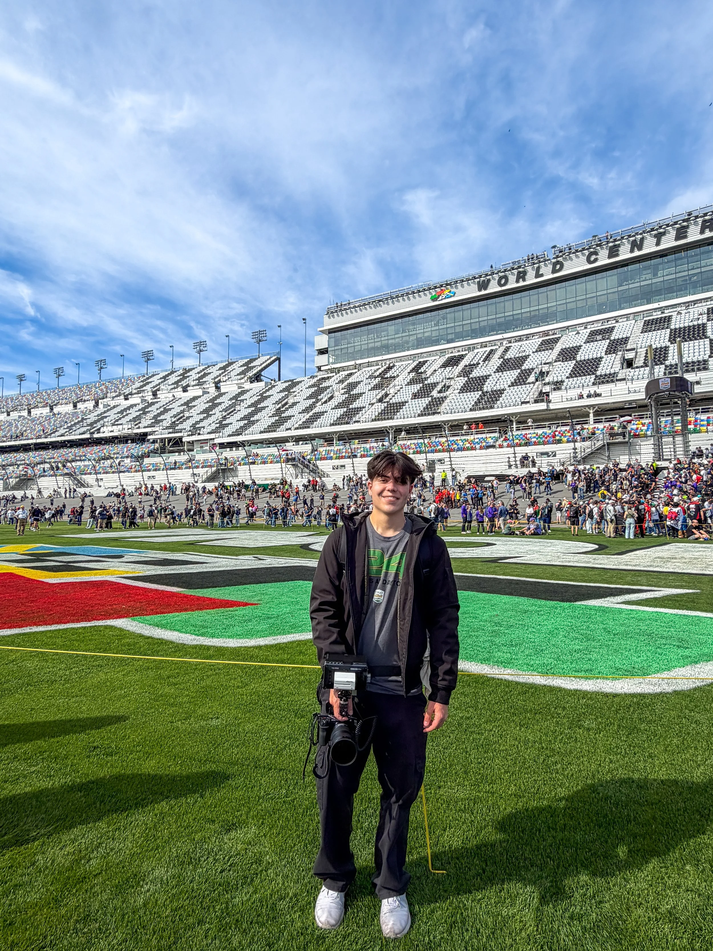 A young man standing on a racetrack at a stadium with a camera in his hand, with the words 'World Center' visible on the building behind him, and a crowd of spectators in the background.