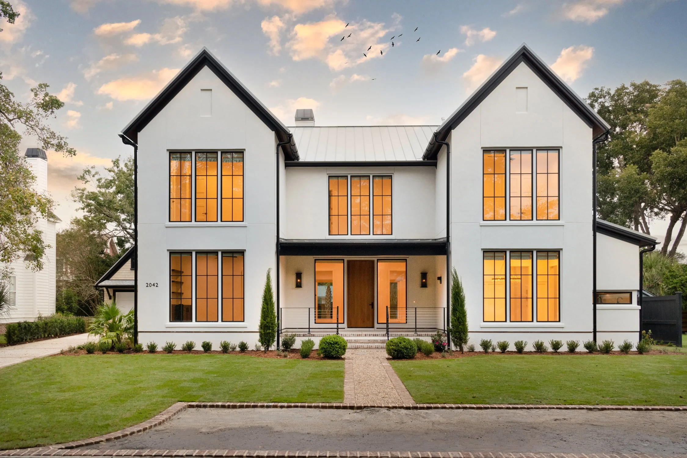 Modern two-story white house with large windows, black trim, and a front porch, surrounded by a well-maintained lawn and garden.