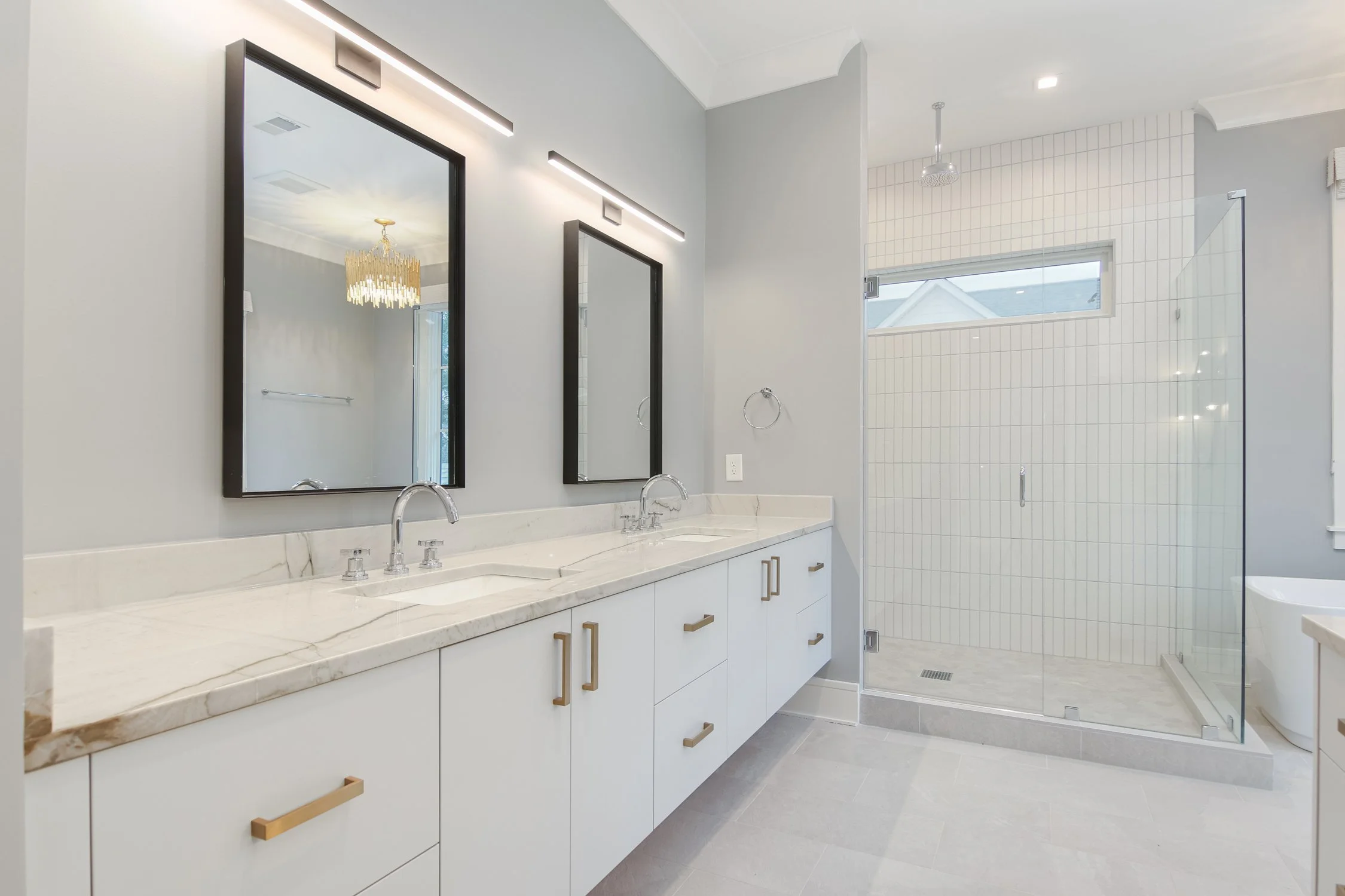 Modern bathroom with double vanity sinks, two mirrors with black frames, and white cabinetry with gold handles. There is a walk-in glass shower with white tile walls and a window.