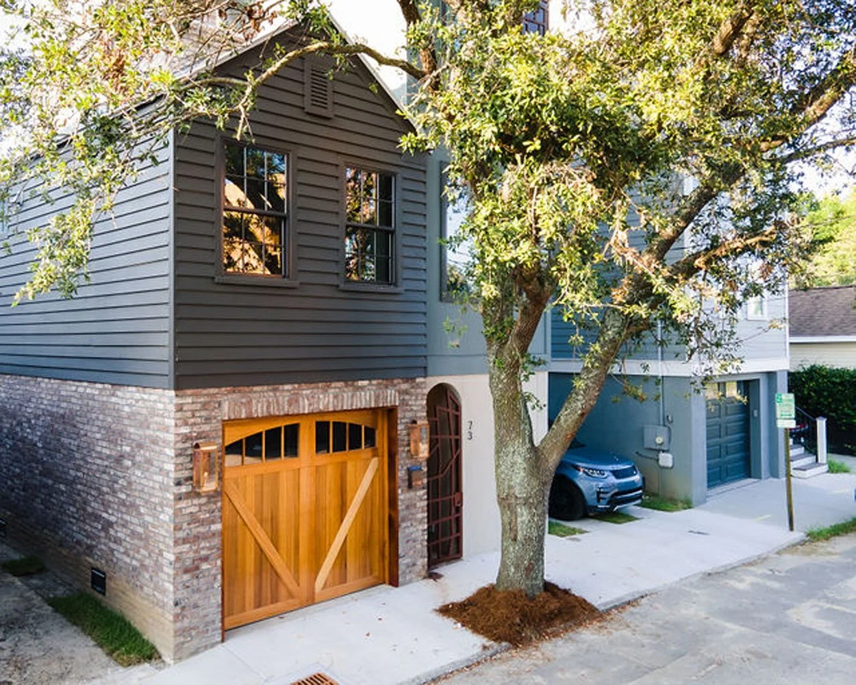 Front view of a two-story house with a brick and gray siding exterior, a wooden garage door, a large tree with green leaves, and a parked car in the driveway.