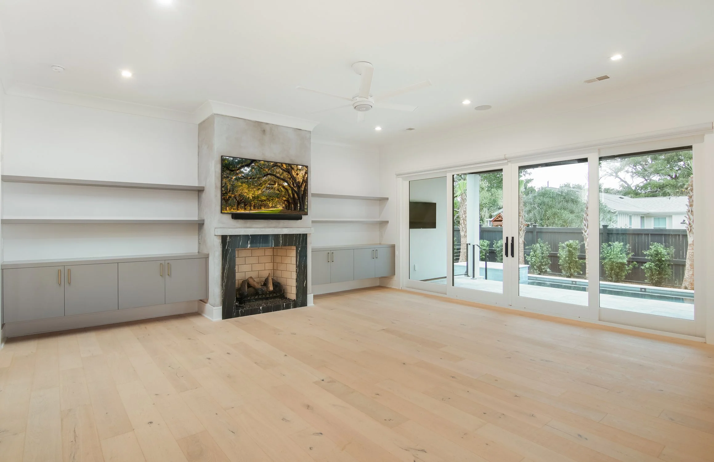 Empty living room with large sliding glass doors, light wooden floors, a fireplace with a TV mounted above, and built-in cabinets and shelves.