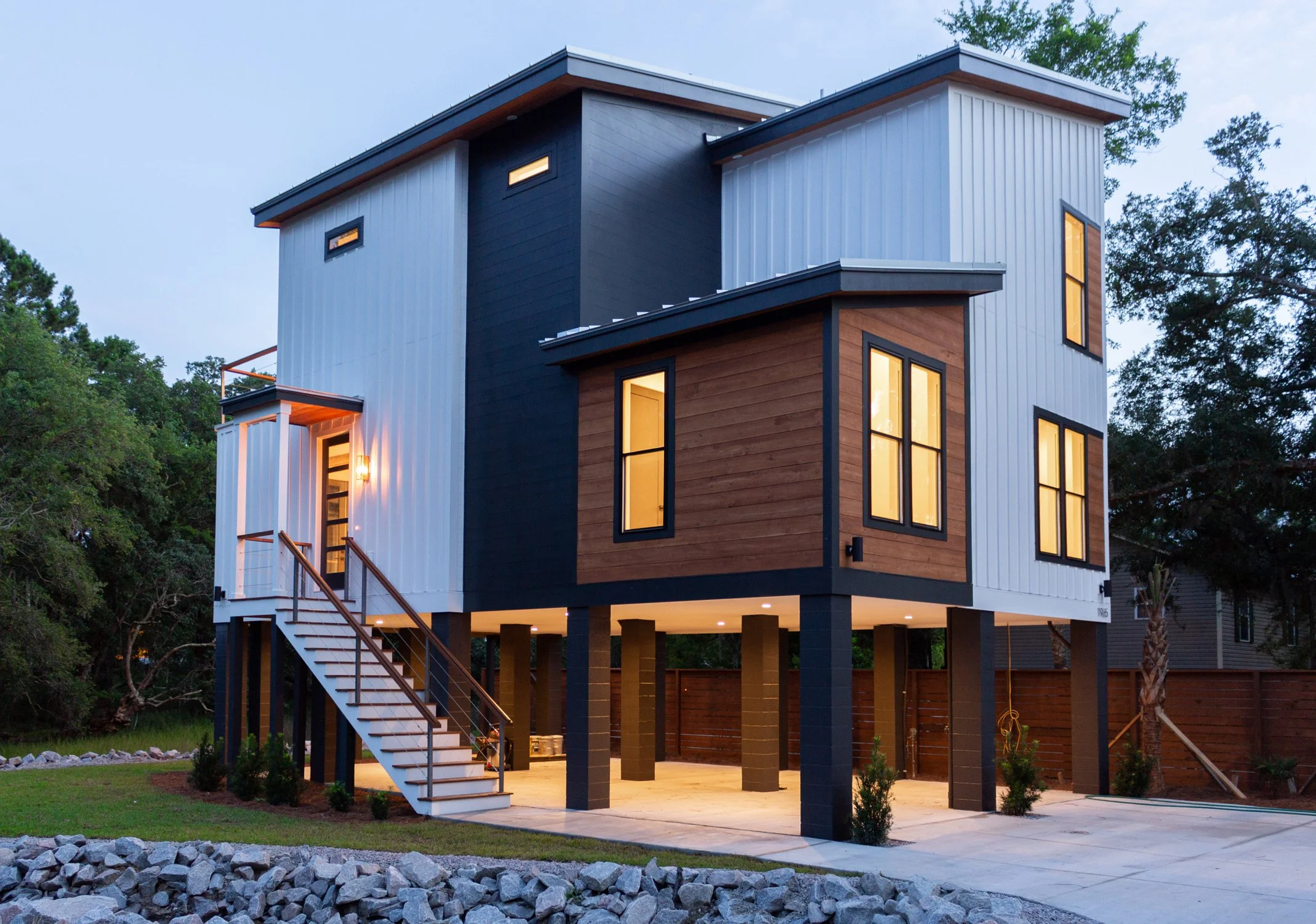 Modern house on stilts with yellow-lit windows, wood and metal exterior, staircase leading to entrance, surrounded by greenery and a stone-bordered yard.