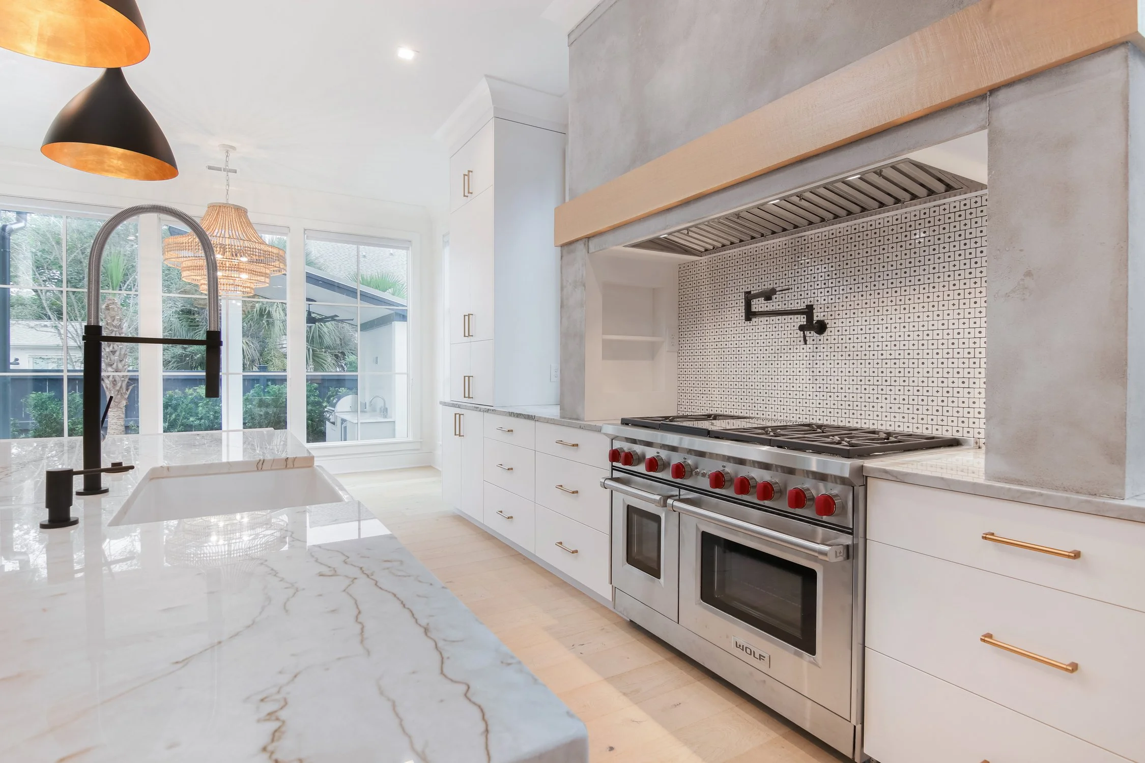 Modern kitchen with white cabinets, marble countertops, a stainless steel double oven, black and gold faucet, and large windows with outdoor view.