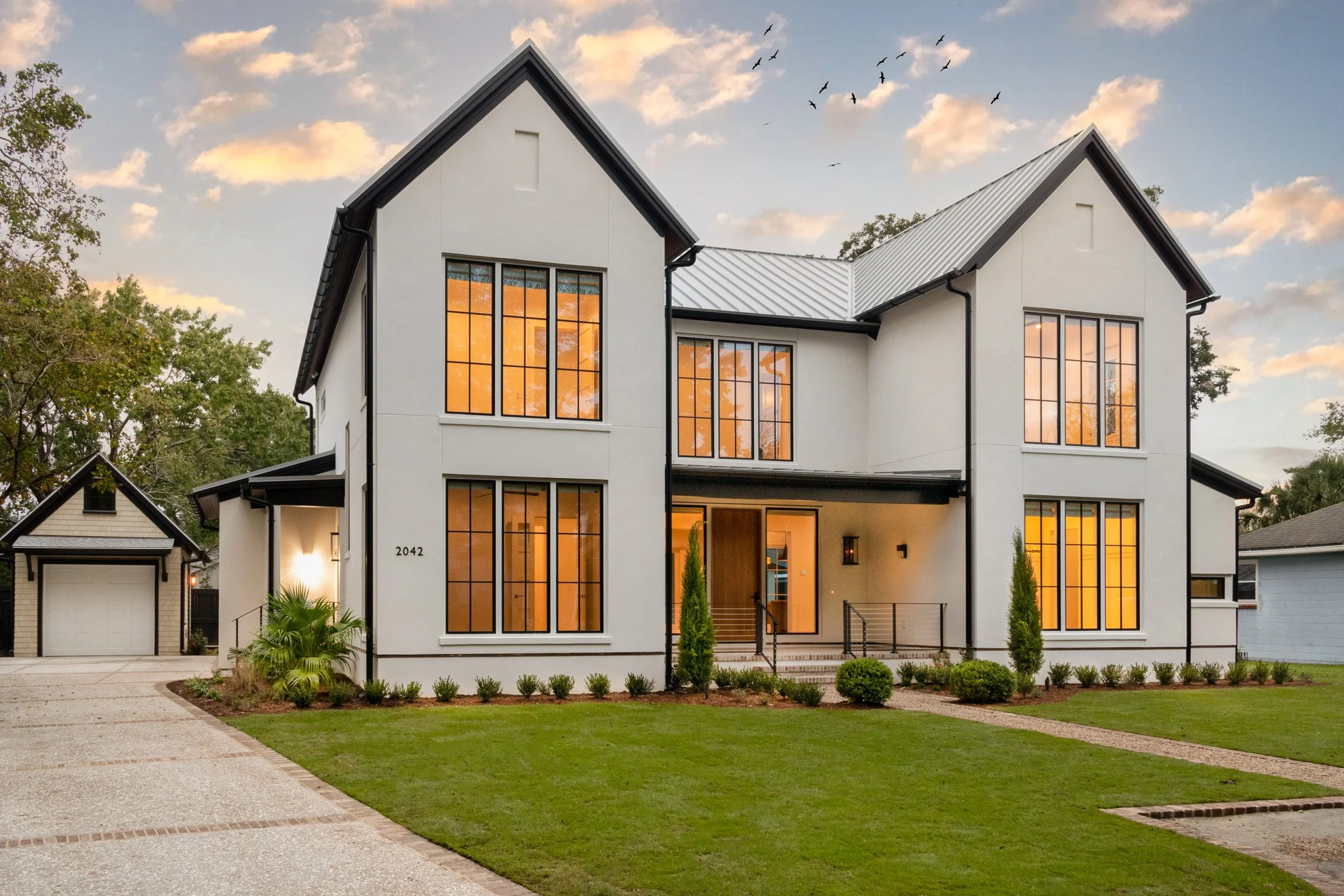 Modern two-story house with white exterior, large windows with warm interior lighting, front yard with green lawn and landscaped plants, driveway leading to a garage, and a sky with clouds and birds flying.