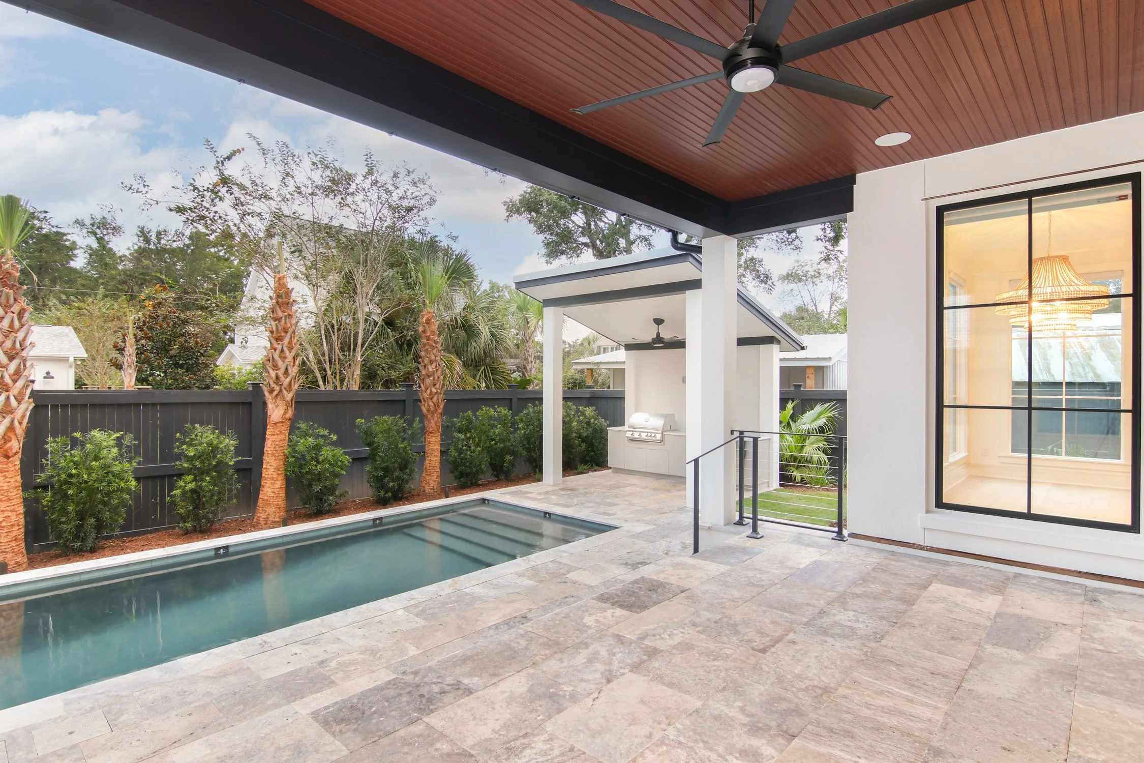 View of a backyard patio with a small swimming pool, outdoor grill, and indoor room visible through large window, with trees and sky in the background.