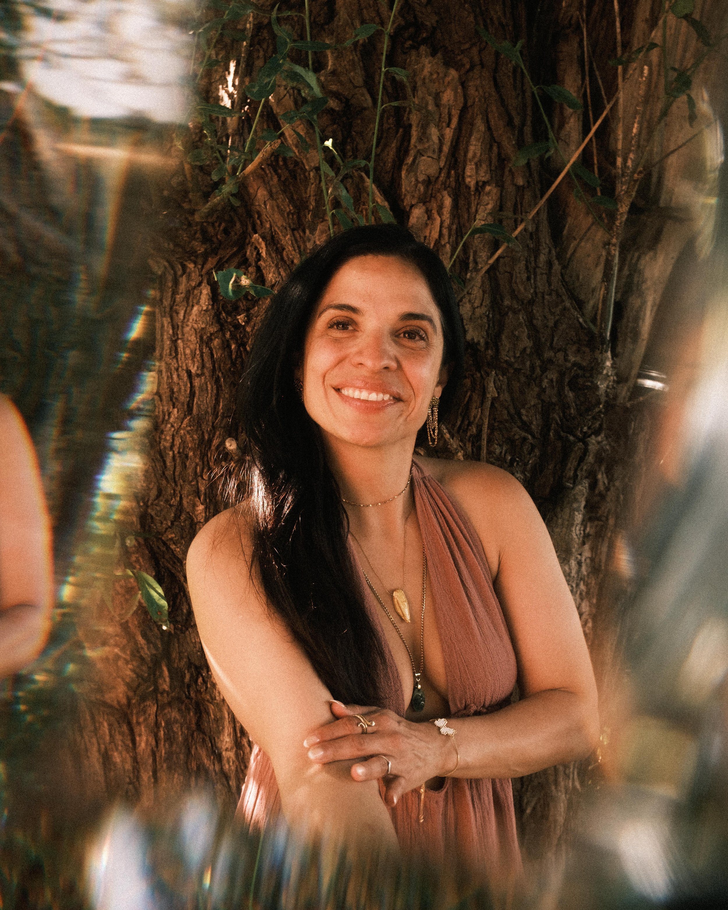 Eine lachende Frau mit schwarzen Haaren, die vor einem großen Baum steht, umgeben von grünen Blättern und buntem Licht. Sie trägt ein rosa Kleid und Schmuck. Die Szene wirkt warm und natürlich.
