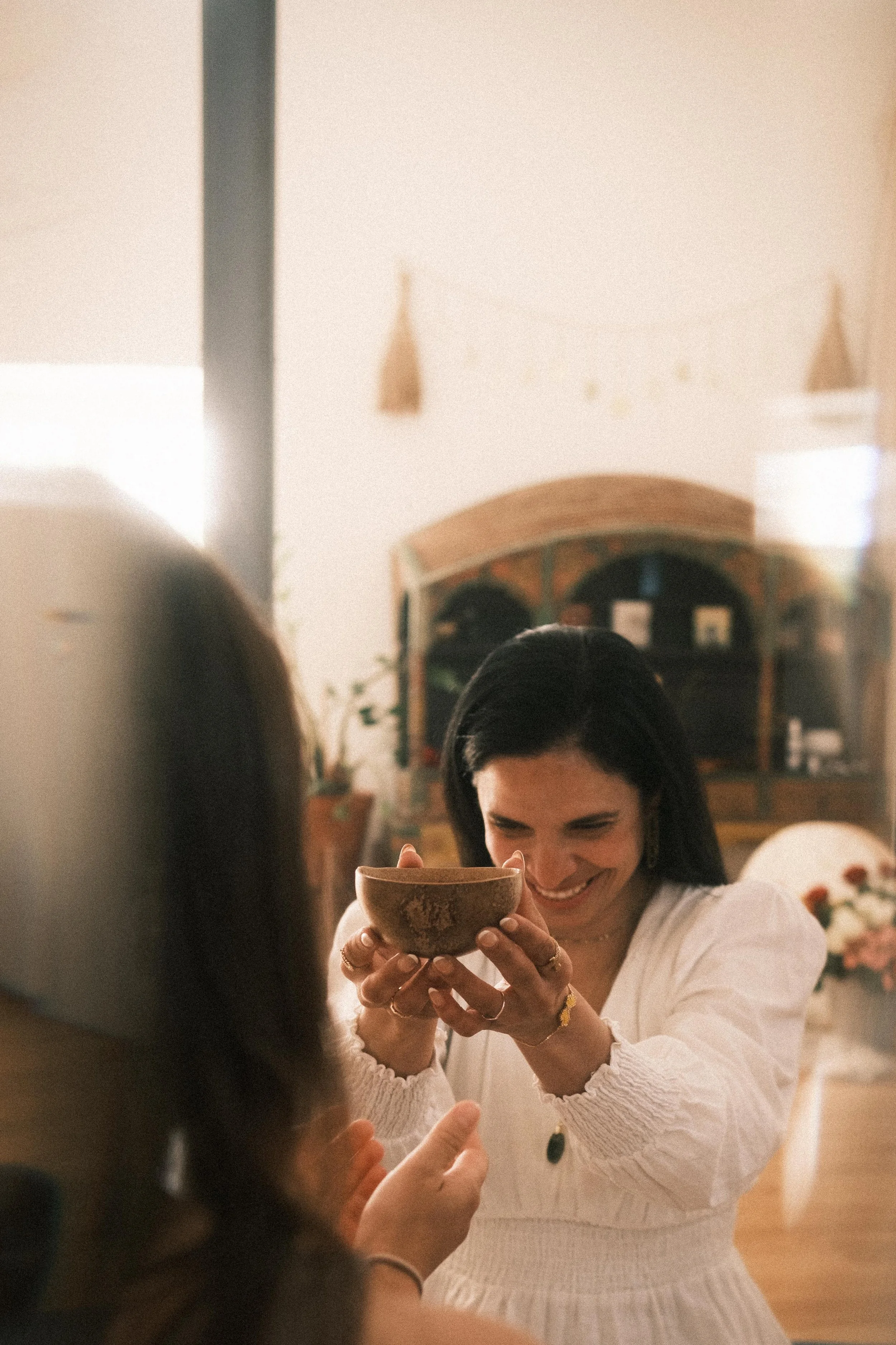 Eine lachende Frau hält eine Schale in die Hand, während sie vor einer anderen Person sitzt, im Hintergrund ein Holzregal und Blumen.