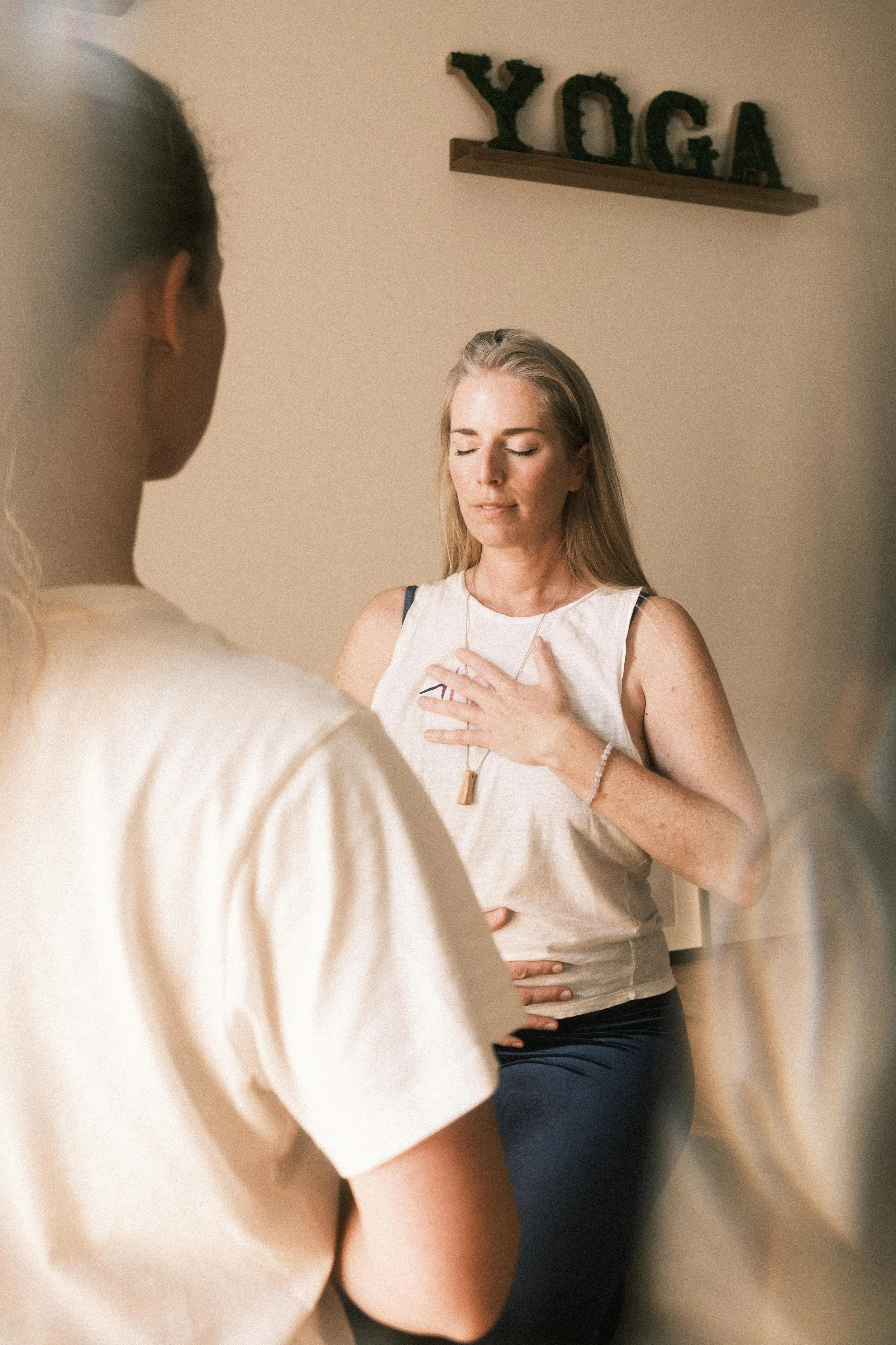 Eine Frau steht meditativ mit geschlossener Augen und Hand auf der Brust, während eine andere Frau im Vordergrund sitzt. Im Hintergrund ist ein Wanddekor mit grünen Buchstaben, die das Wort 'YOGA' bilden.