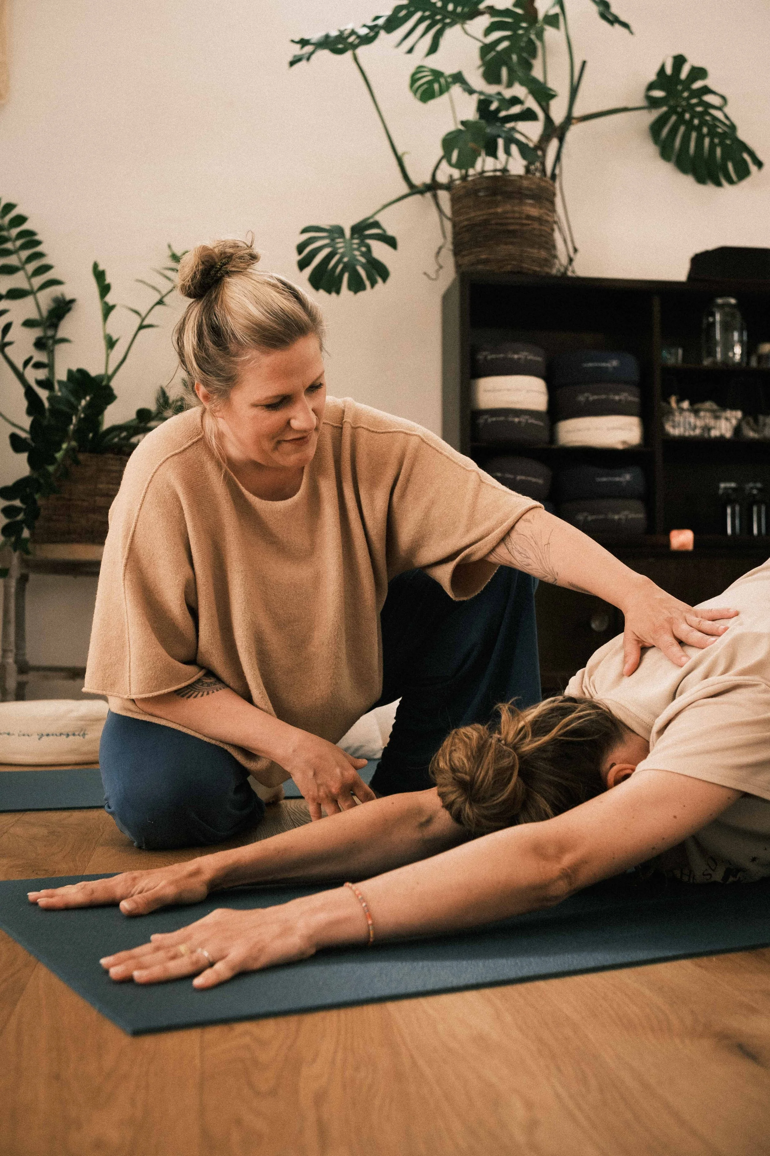 Eine Frau macht Yoga mit einer weiteren Person, die sich nach vorne streckt, in einem Raum mit Pflanzen und Yoga-Accessoires.