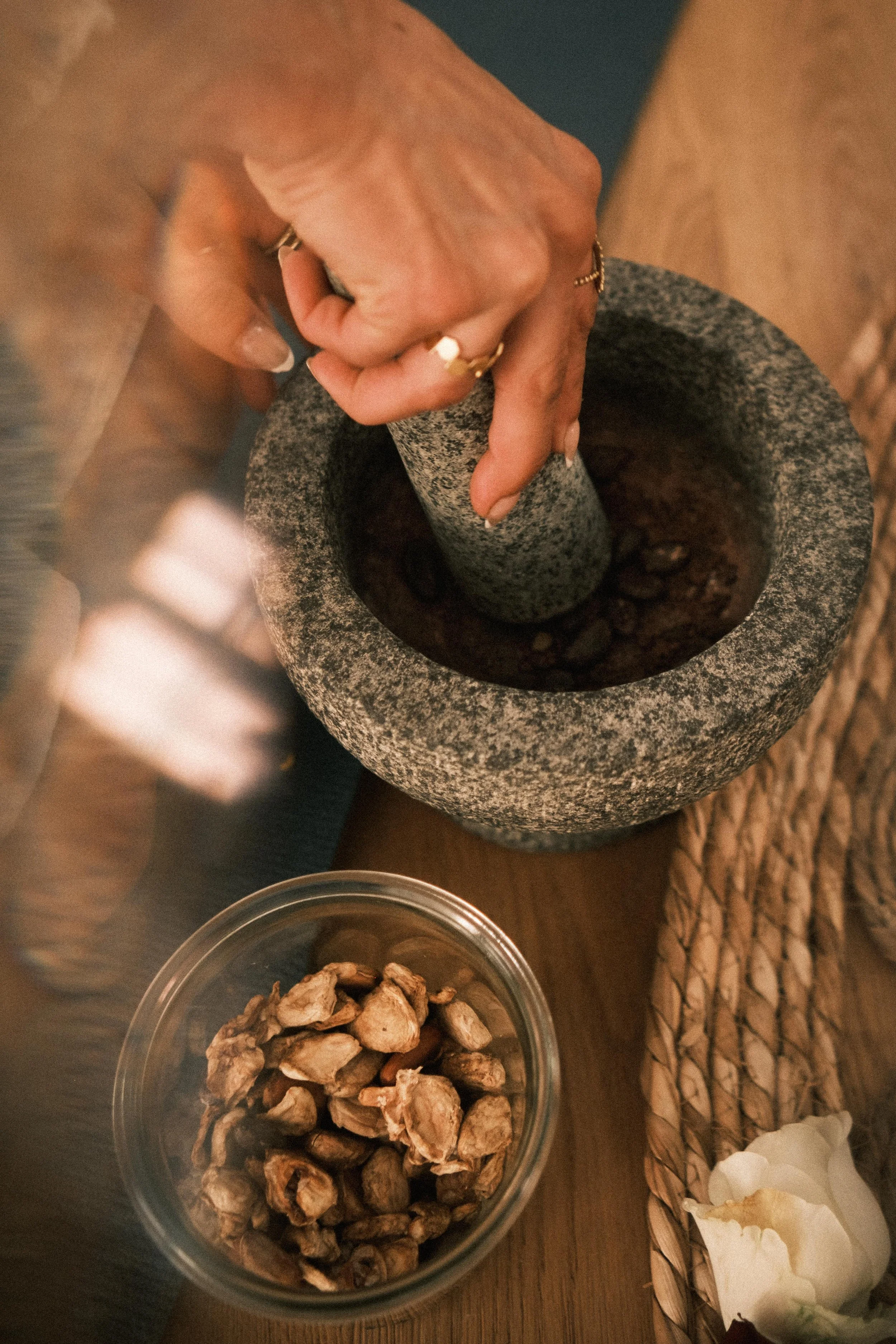 Eine Hand, die mit einem Mörser und Stößel getrocknete Kräuter zermahlt, auf einem Holztisch mit einer Schüssel voller getrockneter Blumen und anderen Gegenständen.