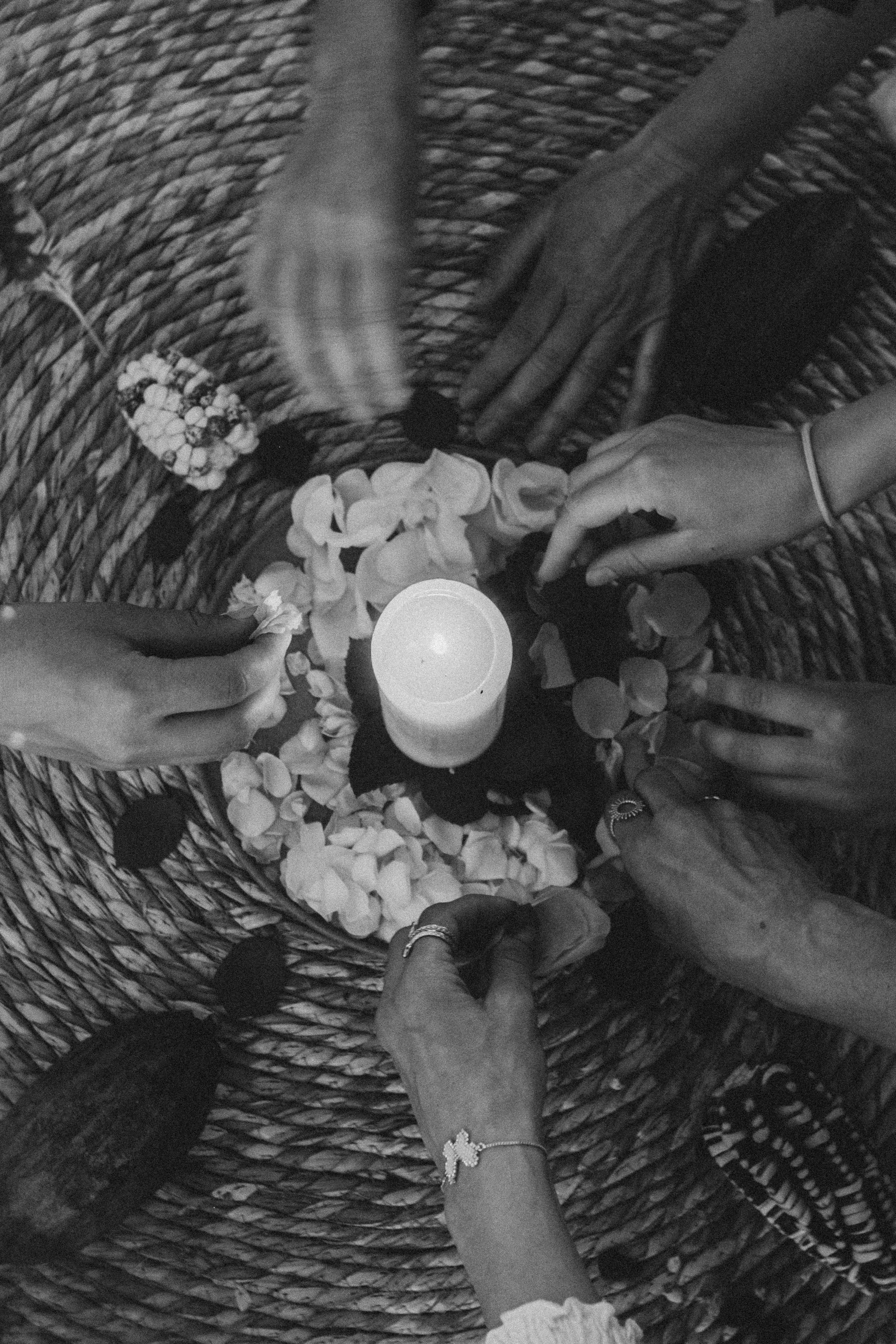 Mehrere Hände, die um eine Kerze herum Blumen und Blütenblätter auf einem geflochtenen Untergrund arrangieren.