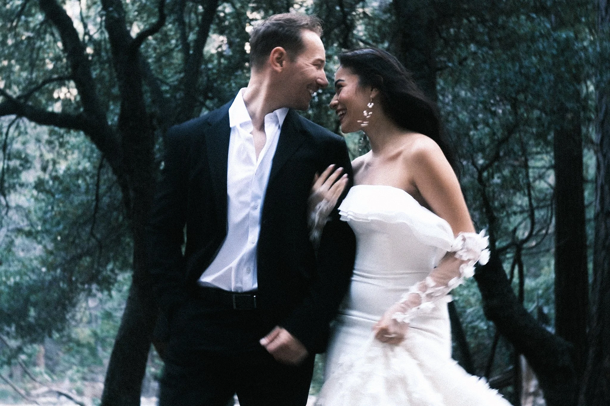 Ein Hochzeitspaar in Anzug und Kleid spaziert durch den Wald und lacht.