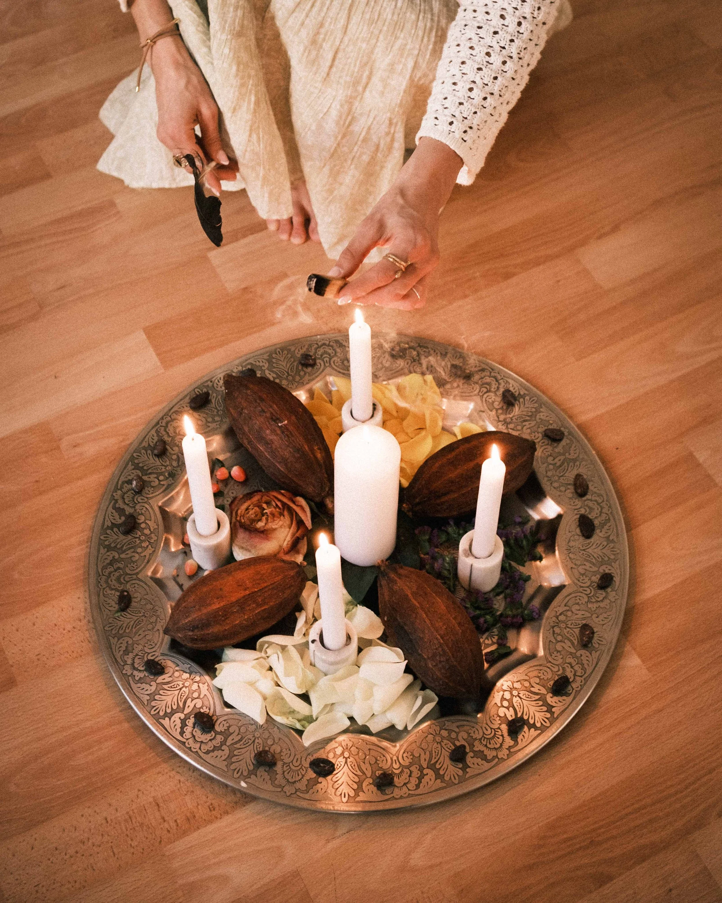 Eine Person zündet Kerzen auf einem dekorativen, runden silbernen Tablett mit Kräutern, Blumen und Kakaobohnen. Die Person trägt mehrere Ringe und Schmuck.