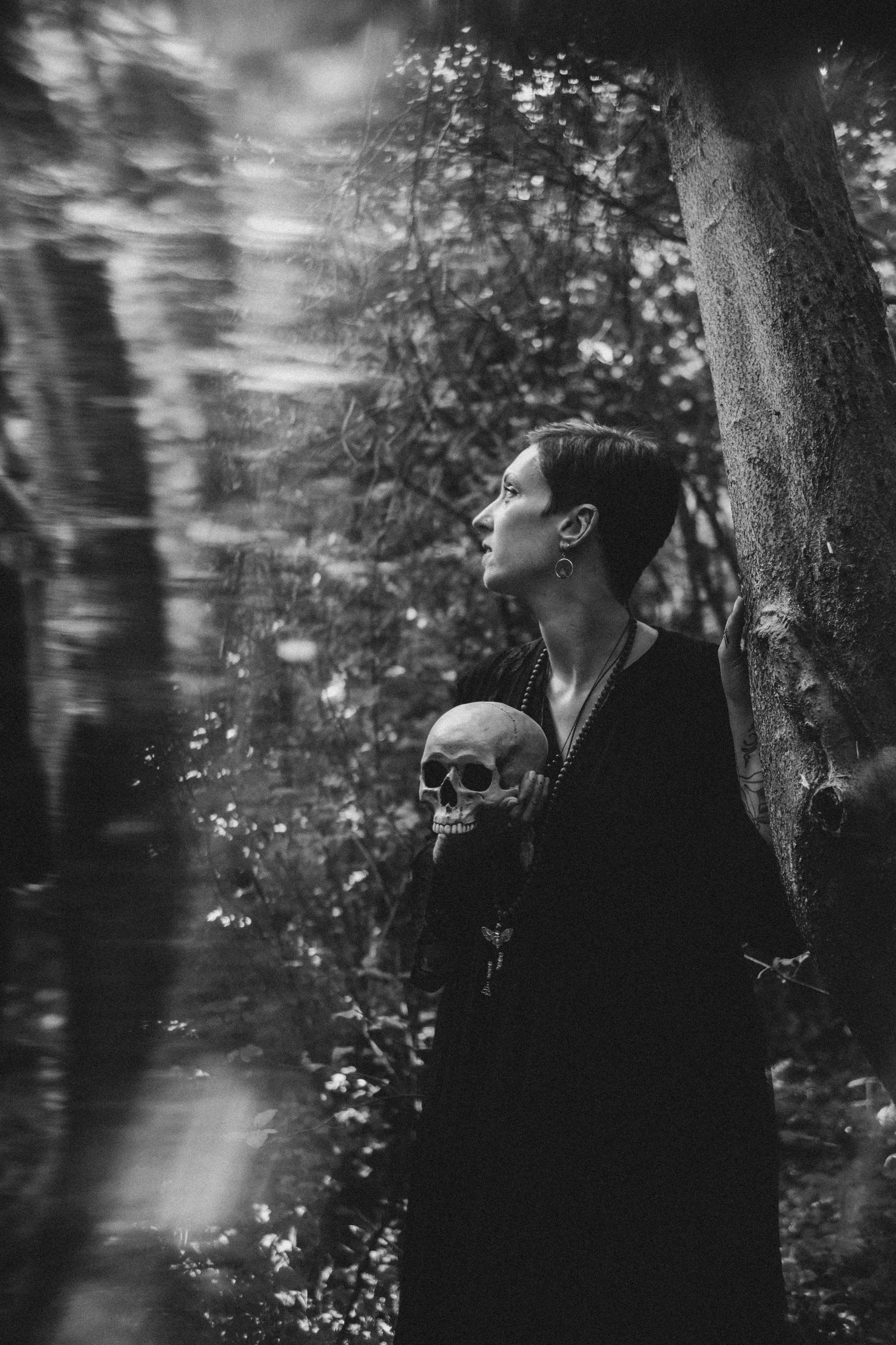 Eine Frau mit kurzen Haaren, dunklem Kleid, Ohrringen und Halskette, steht im Wald, hält einen Totenschädel in der Hand und lehnt sich an einen Baum. Das Bild ist schwarz-weiß.