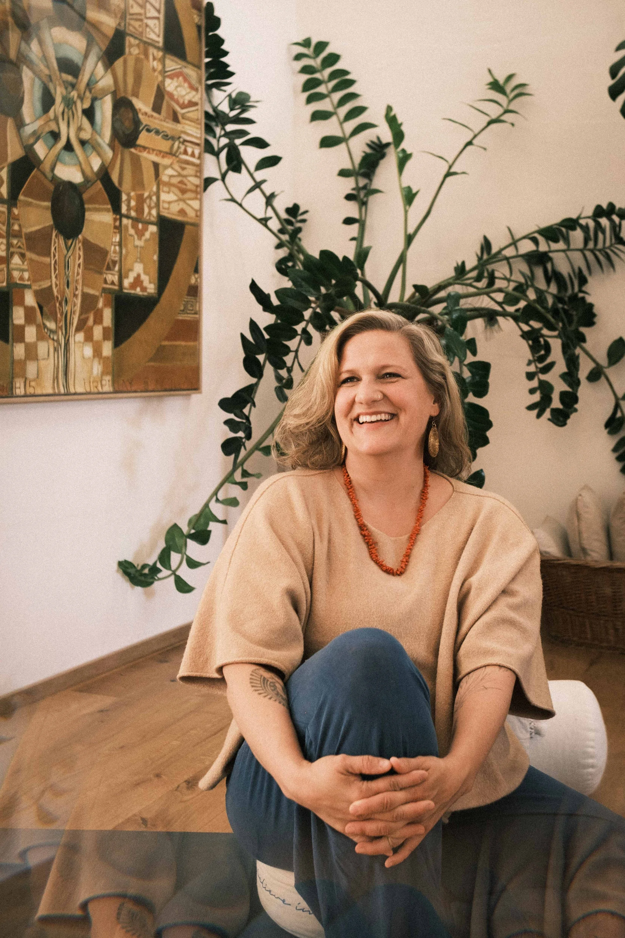 Eine lachende Frau sitzt auf dem Boden in einem Raum mit Holzboden, vor einer großen Pflanze und einem Gemälde an der Wand.