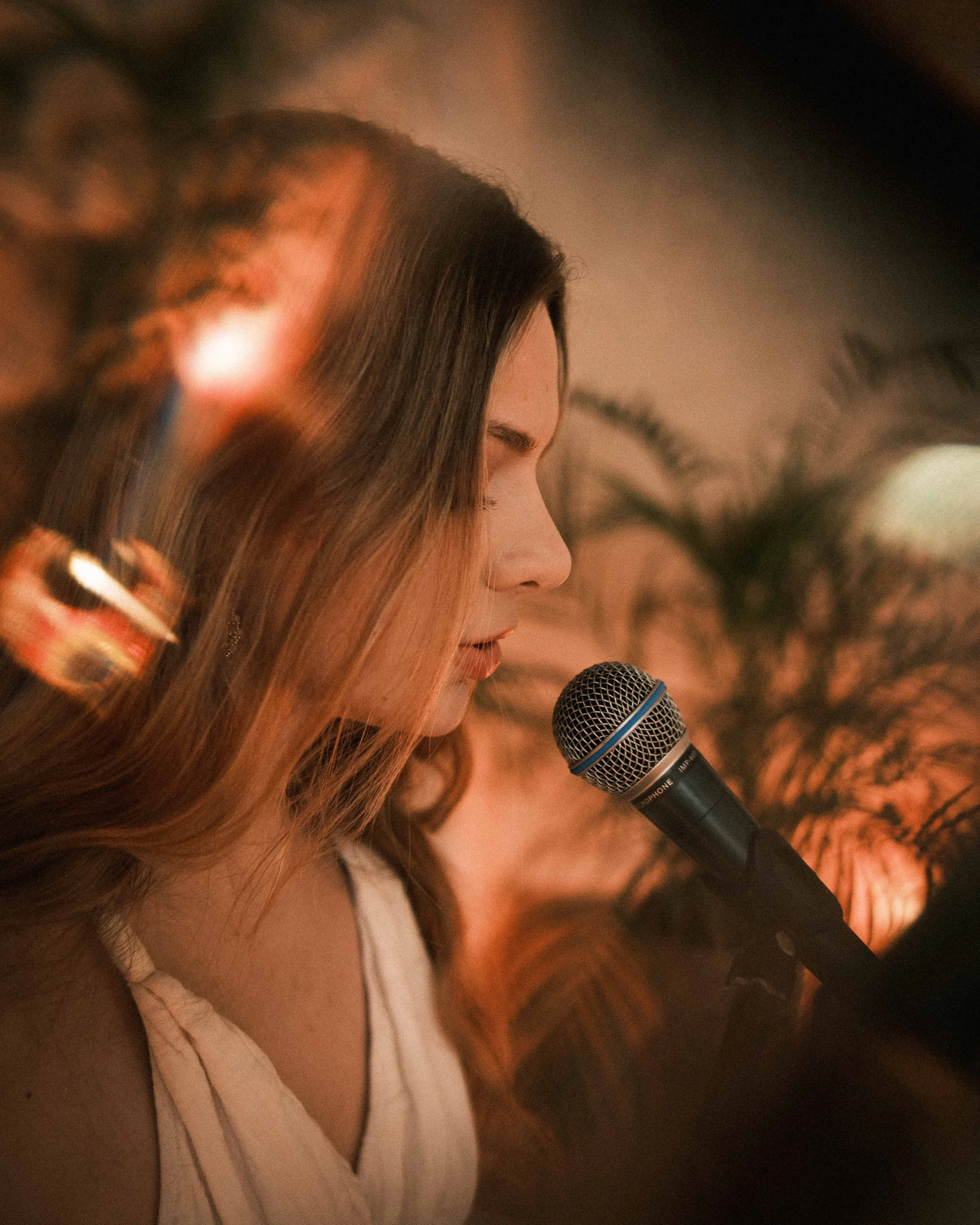 Junge Frau mit braunen Haaren singt in ein Mikrofon, im Hintergrund eine Palme.