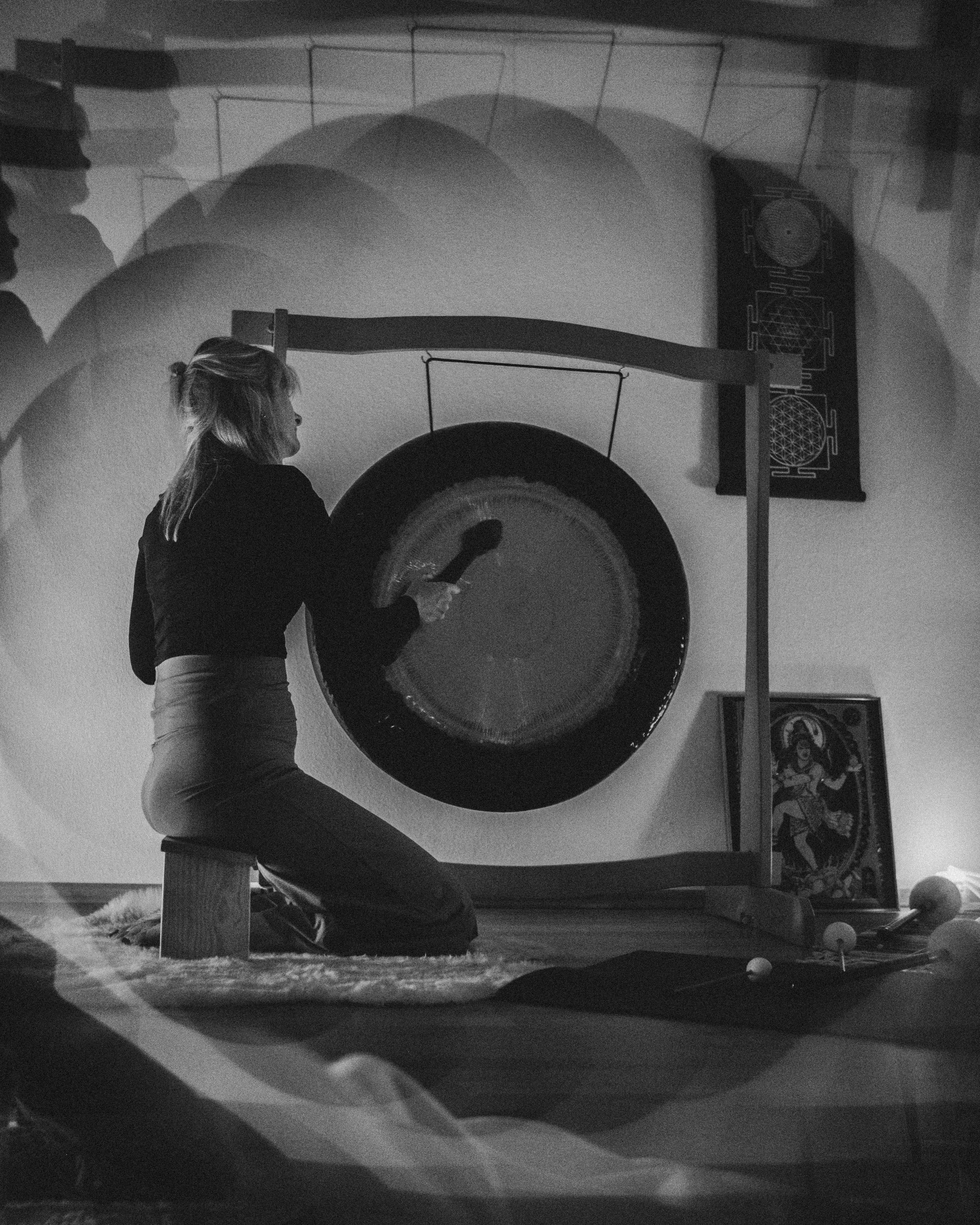 Frau schlägt mit einem Schlägel auf eine große gongähnliche Trommel. Die Szene ist in Schwarzweiß, und im Hintergrund sind schattierte erotische Kunstwerke. Die Frau sitzt auf einem Schemel auf Teppichboden.