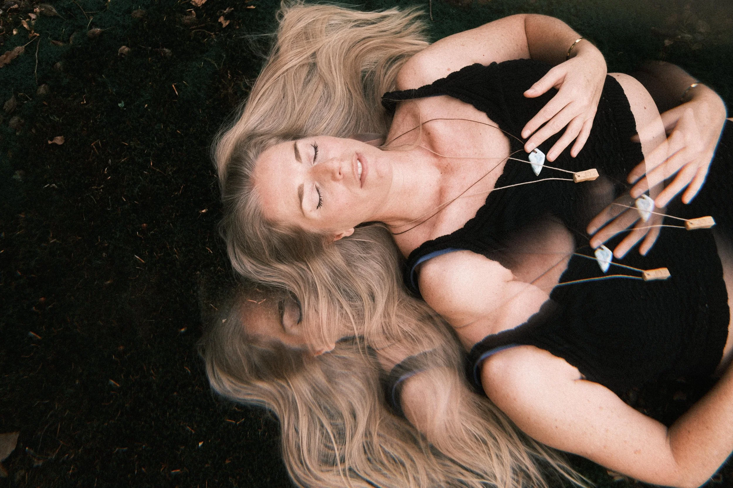 Eine Frau mit langen blonden Haaren liegt auf Waldboden, die Augen geschlossen, mit einer Halskette. Sie trägt ein schwarzes, ärmelloses Kleid und wirkt entspannt.