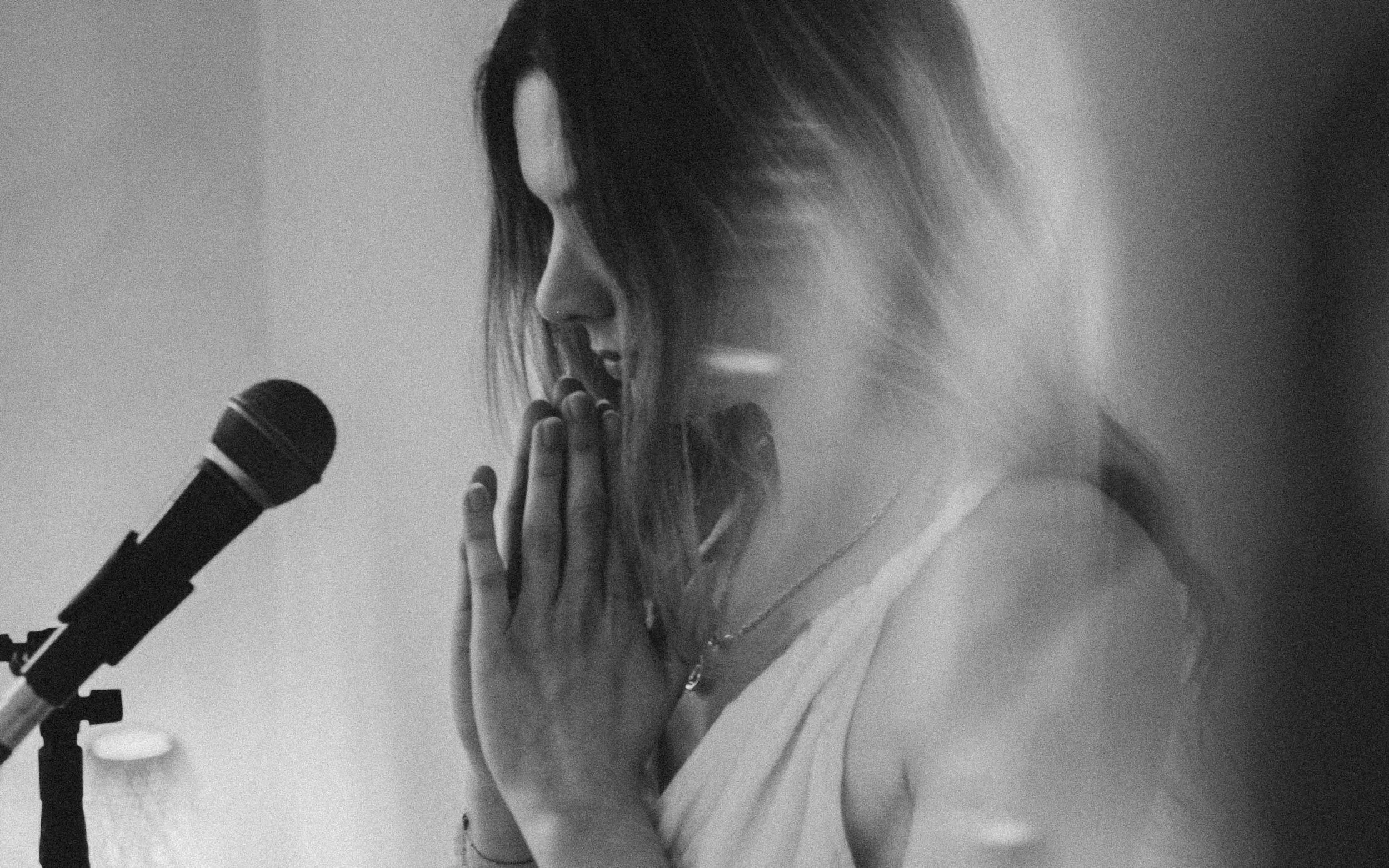 Schwarz-weiß-Foto eines Sängerin, die mit gefalteten Händen vor einem Mikrofon sitzt, den Blick nach unten gerichtet.