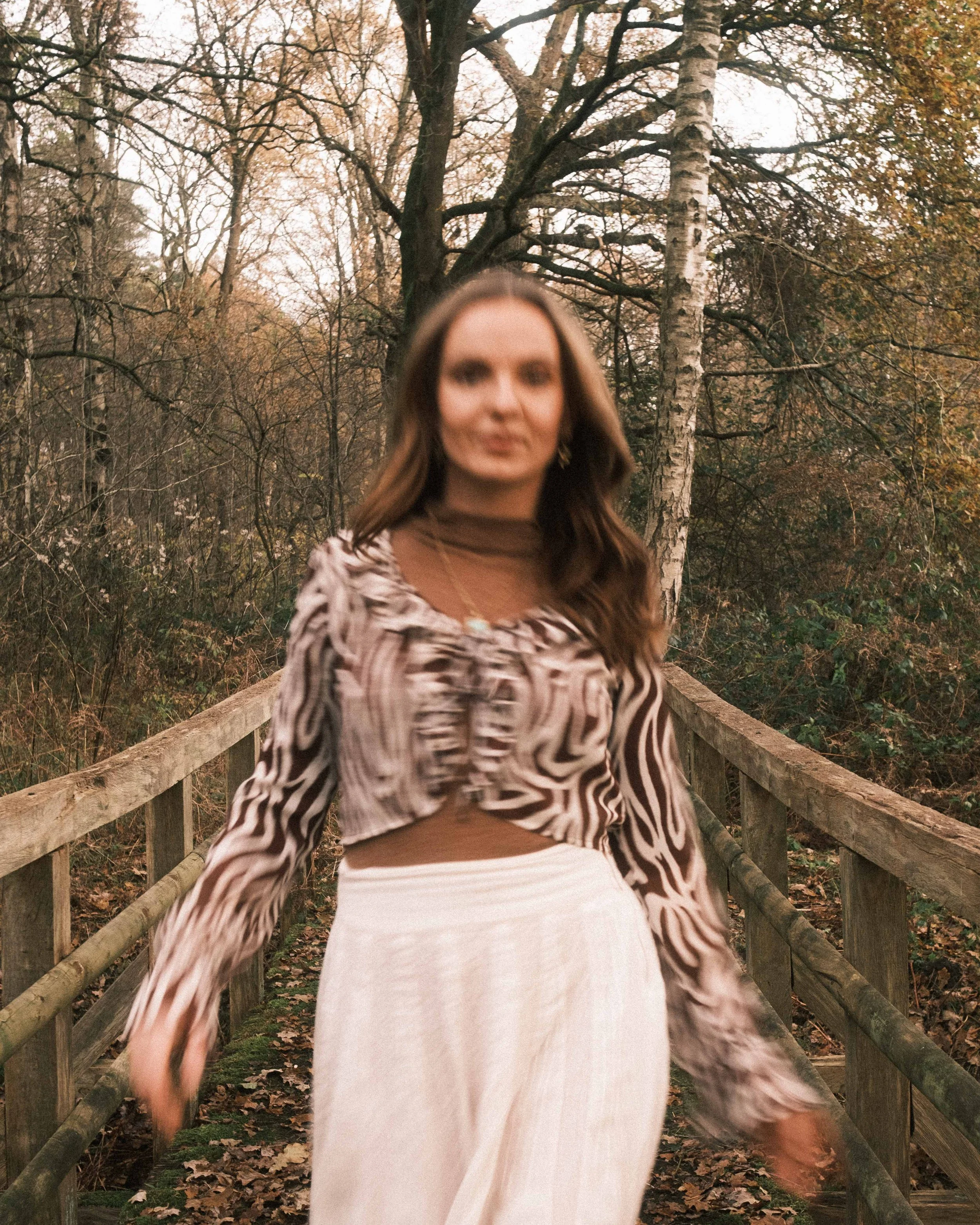 Frau mit langem braunem Haar, Zebra-Muster-Bluse, brauner Turtleneck, weißem Rock, auf einer Holzbrücke im Wald im Herbst, unscharf im Hintergrund.