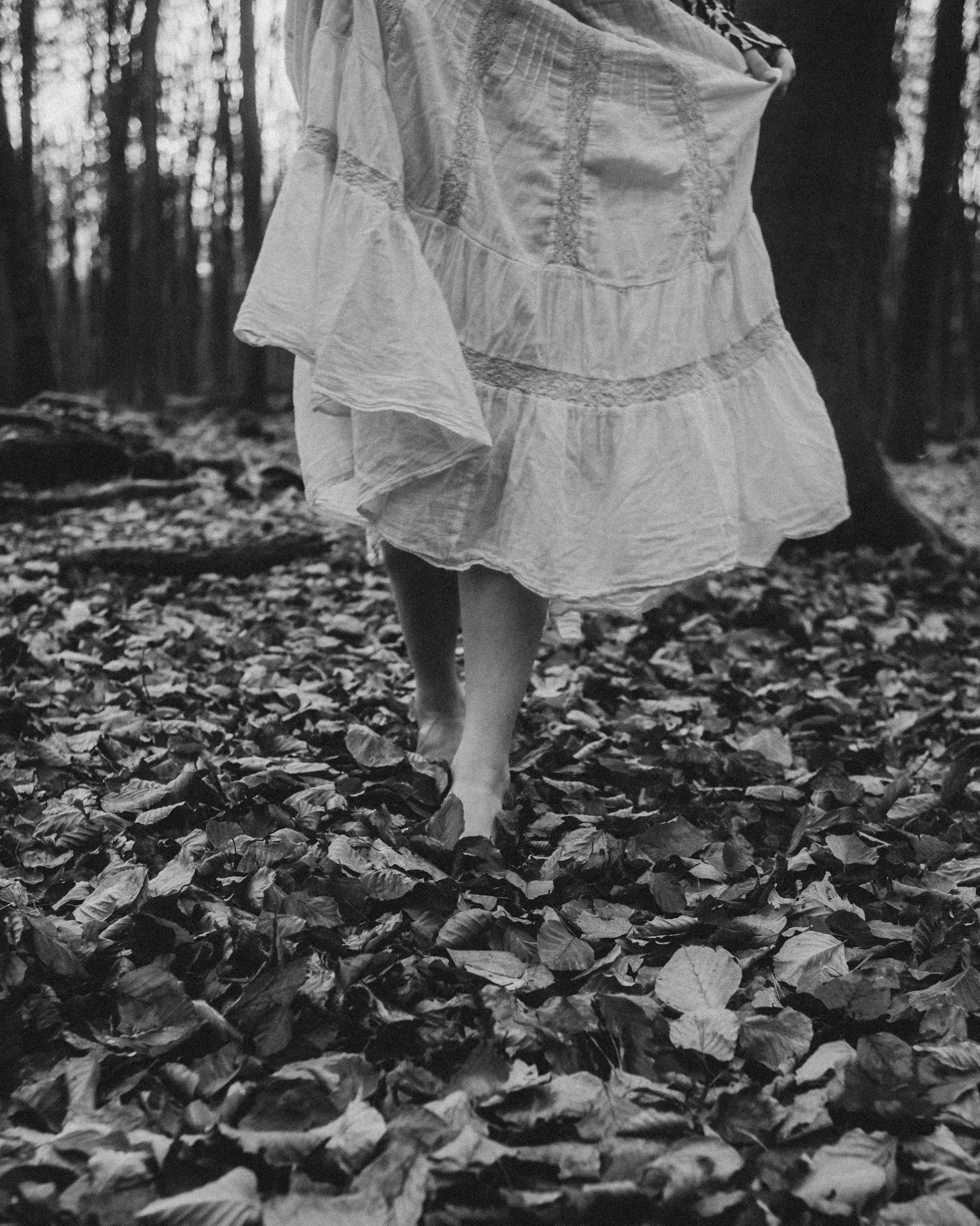 Eine Person ist in einem Wald unterwegs, trägt ein fließendes Kleid und ist barfuß auf einem mit Blättern bedeckten Boden. Das Bild ist in Schwarz-Weiß gehalten.