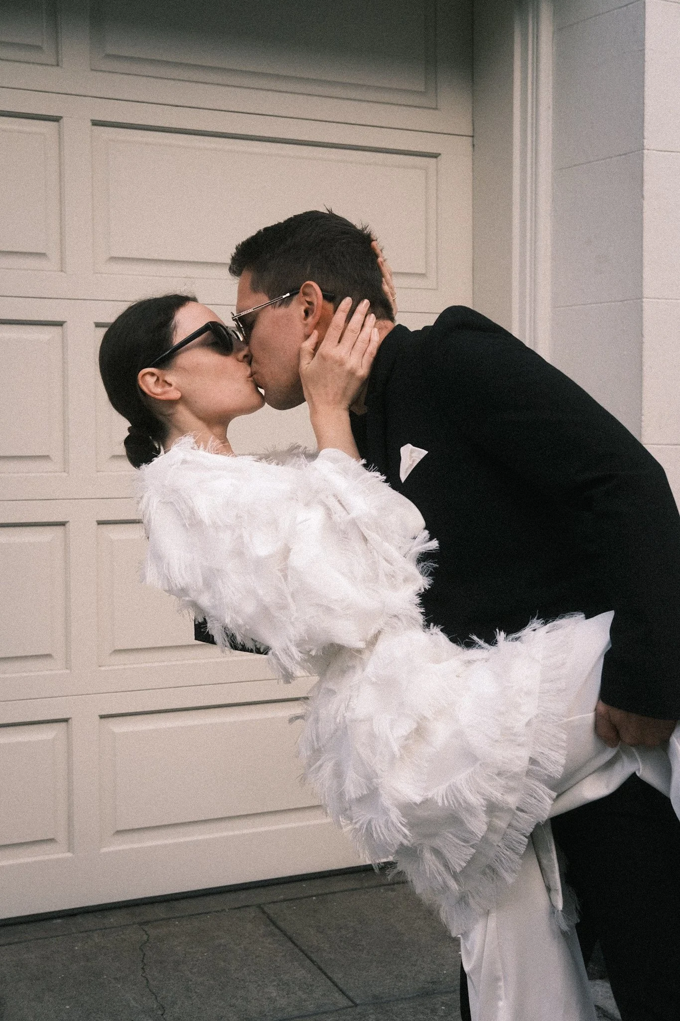 Ein Paar küsst sich, die Frau trägt einen weißen Designer-Anzug mit Sakko in Feder-Optik und eine Sonnenbrille, der Mann trägt einen schwarzen Anzug mit weißem Taschentuch in der Brusttasche.