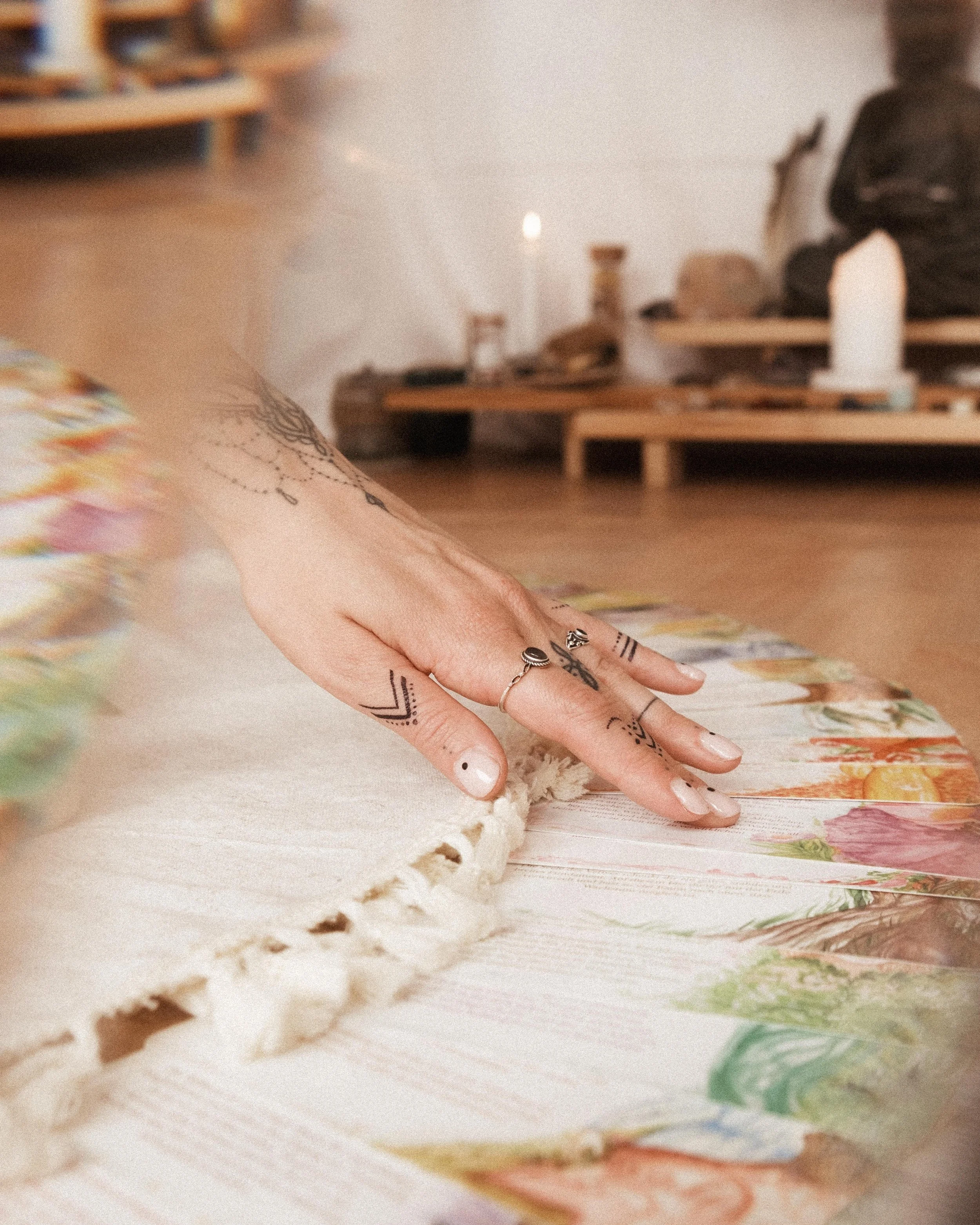Hand mit Tattoos, die eine bunte Karte auf einem Tisch berührt, im Hintergrund sind Kerzen und Dekorationen sichtbar