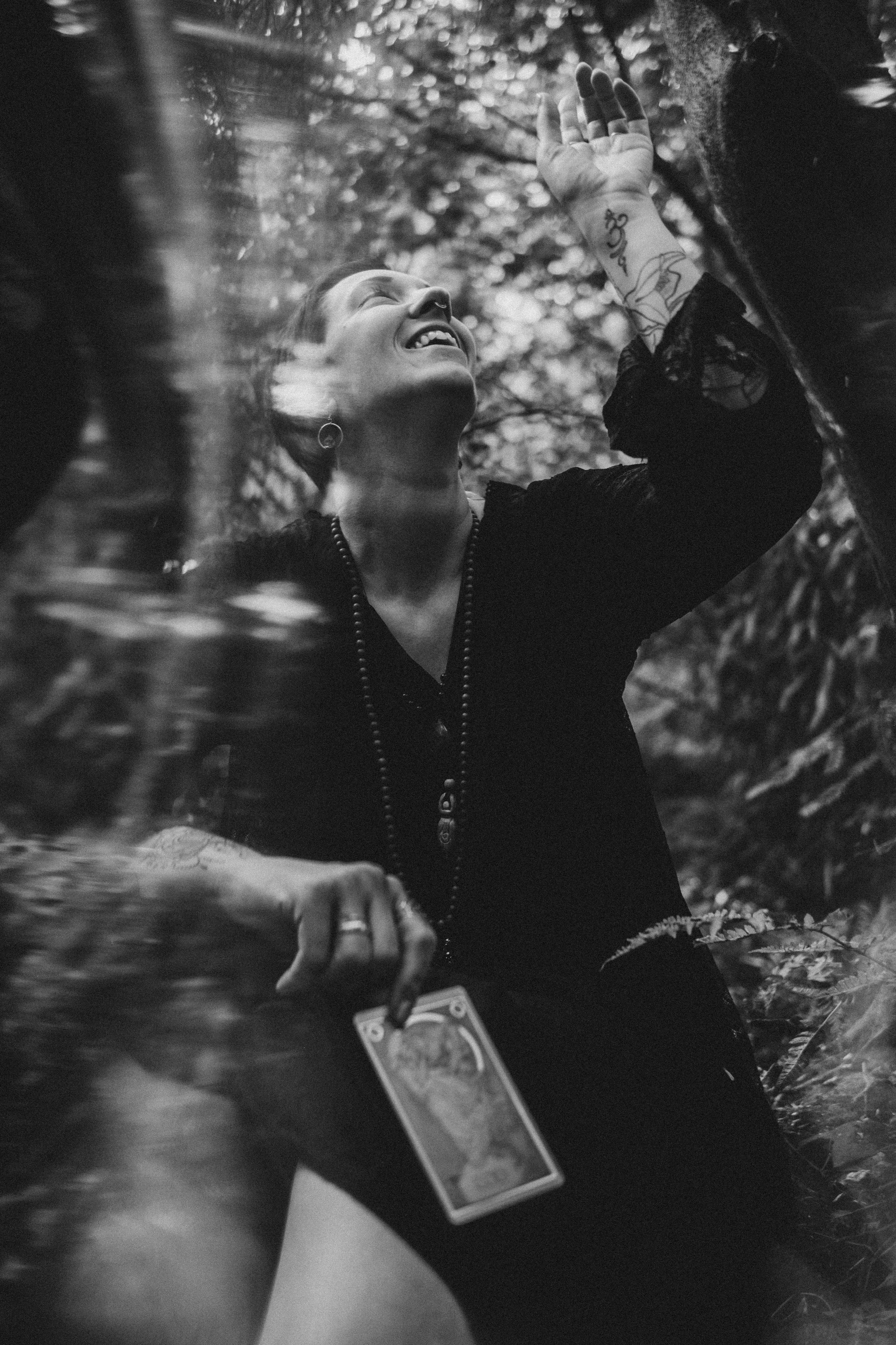 Schwarz-Weiß-Foto einer Person mit Tattoos, die im Wald unter Bäumen steht, lächelnd nach oben schaut, eine Hand hebt und eine Tarotkarte hält.