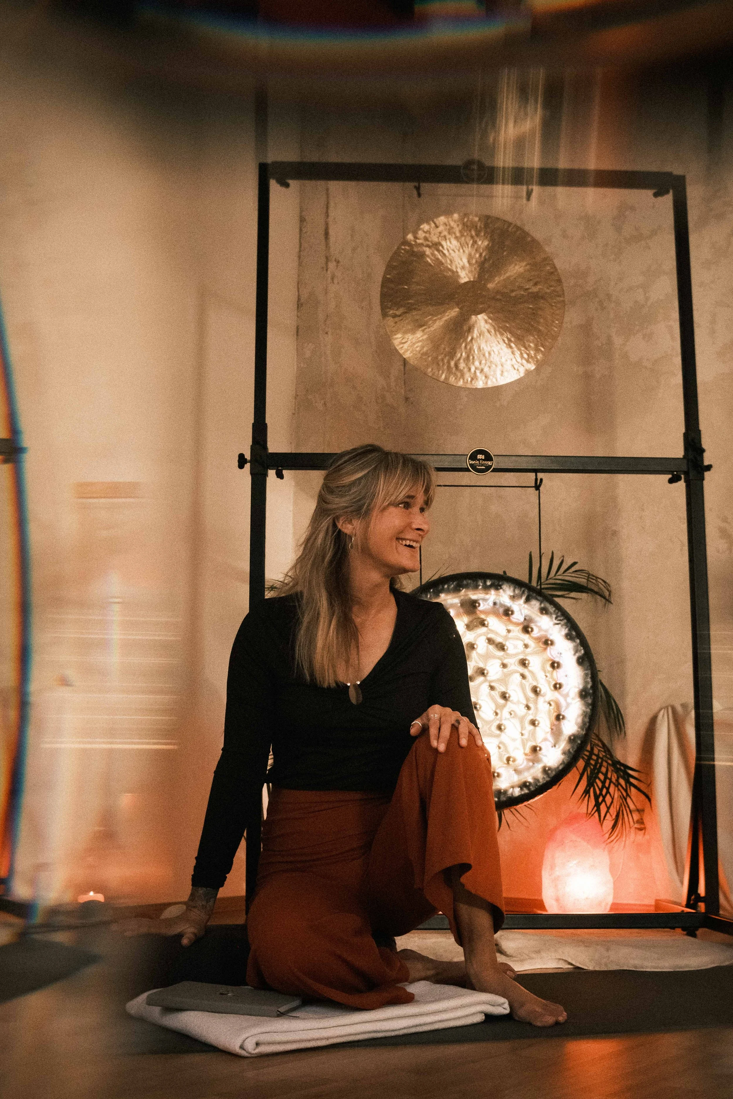 Eine lachende Frau sitzt im Yoga-Kurs auf einer Matte, im Hintergrund therapeutische Muscheln und eine Decke, in einer gemütlichen, warm beleuchteten Umgebung.