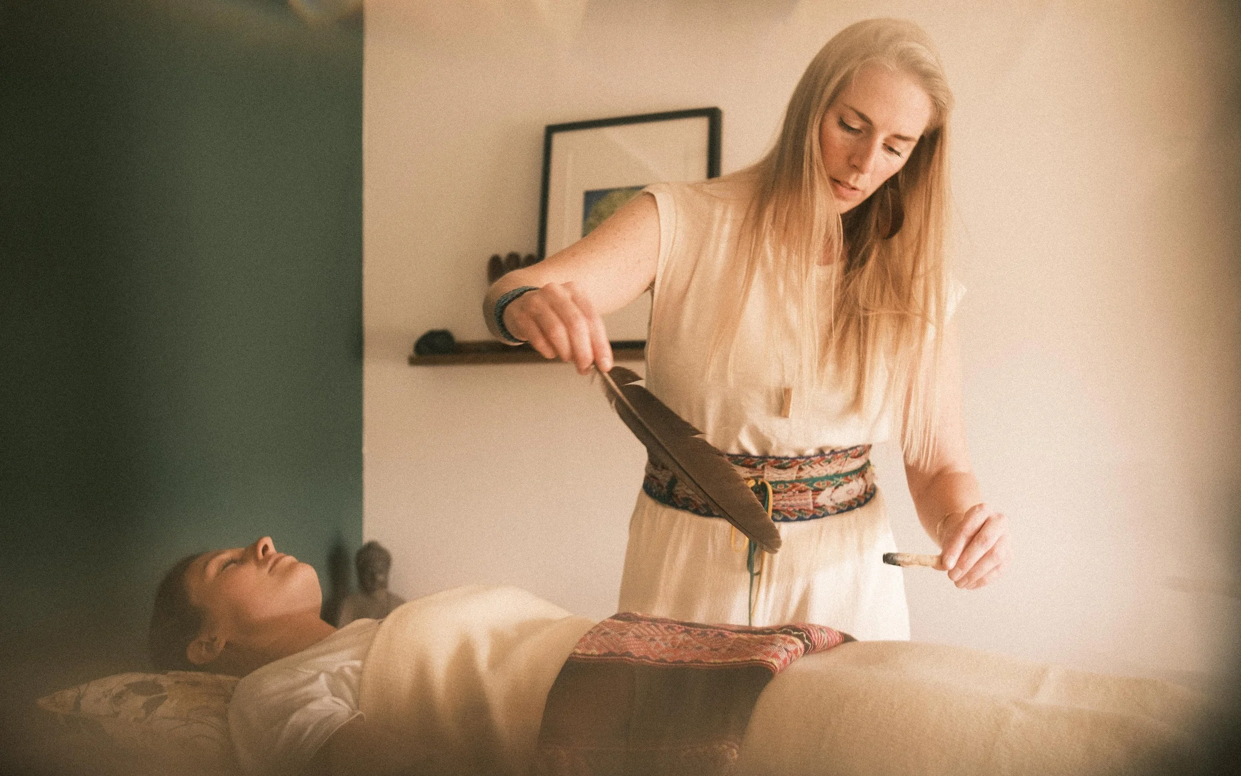 Eine Frau führt eine schamanische Zeremonie durch, indem sie mit einer Feder und Rauch über eine liegende Person schwenkt, in einem Raum mit neutraler Wandfarbe.