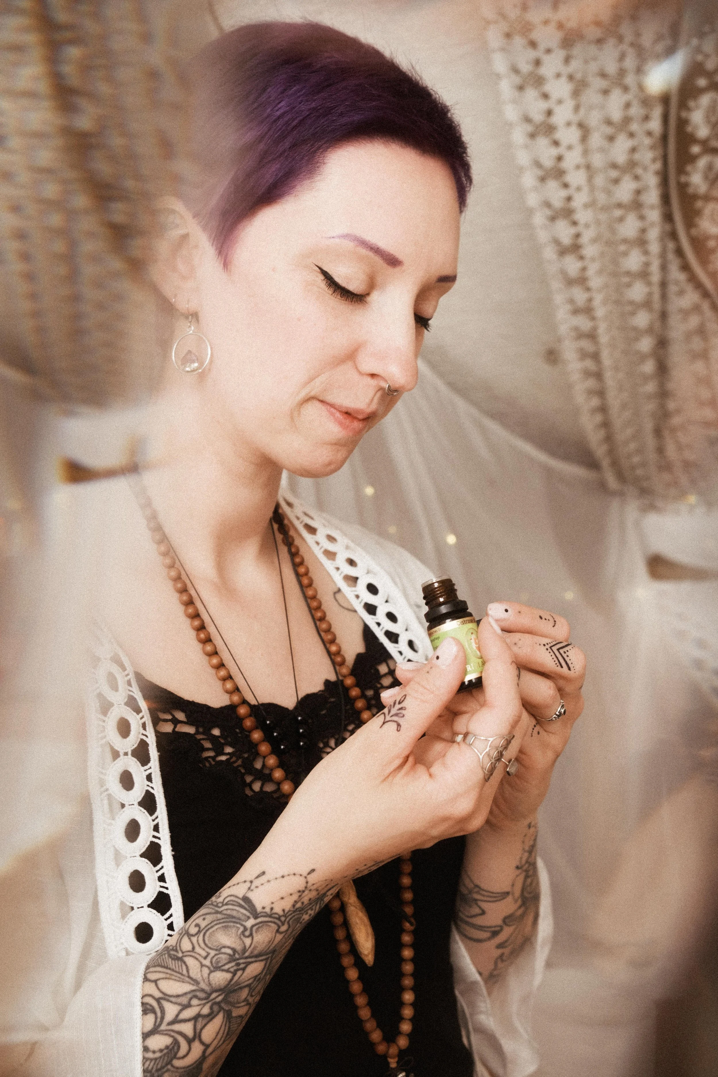 Eine junge Frau mit Tattoos, die ein Fläschchen ätherisches Öl hält und daran riecht, trägt mehrere Halsketten, Ohrschmuck und einen weißen, lochgeschnittenen Mantel über einem schwarzen Top.