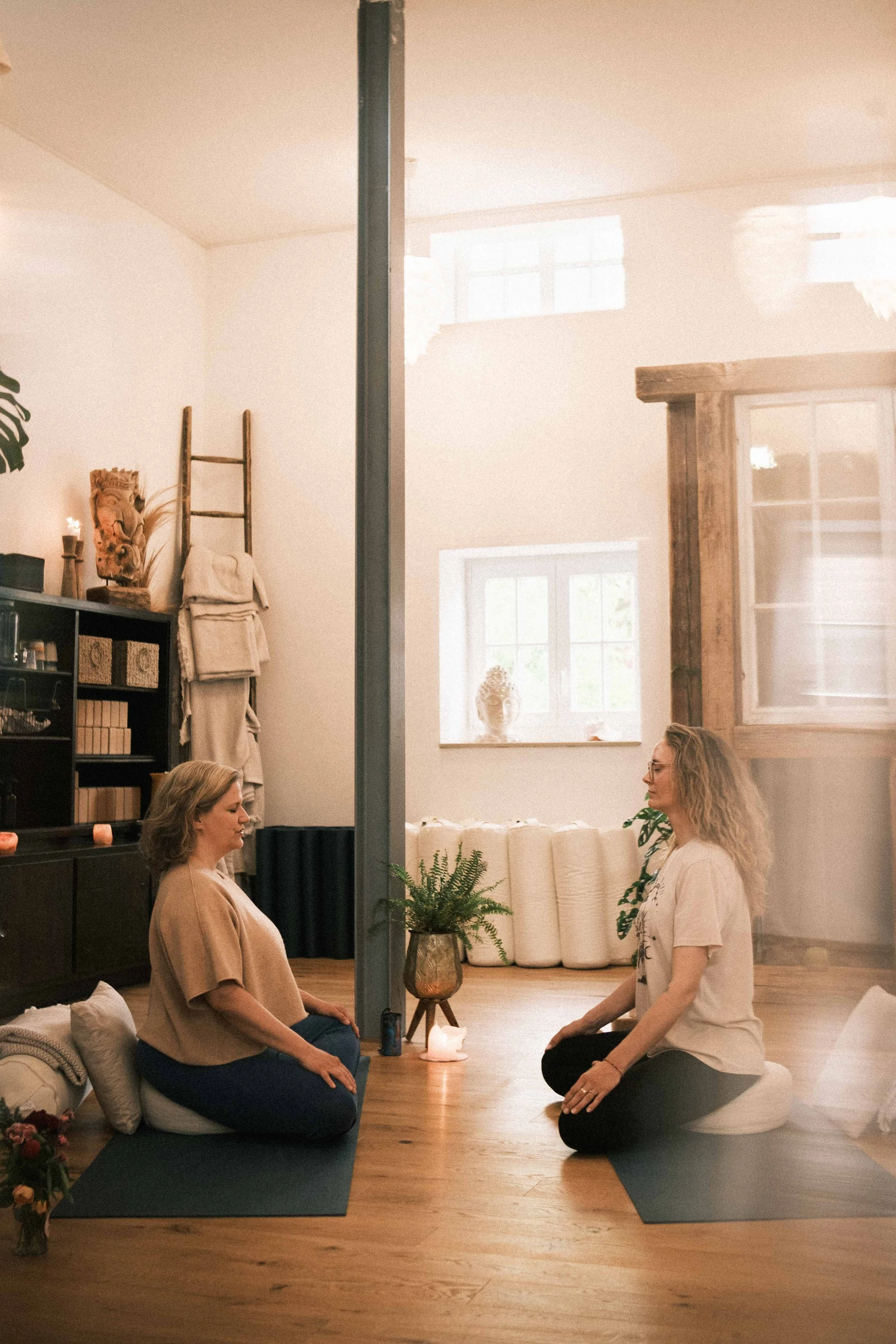 Zwei Frauen beim Yoga oder Meditation, sitzend auf Yogamatten im Raum, umgeben von Dekoration und Pflanzen, in entspannter Atmosphäre.