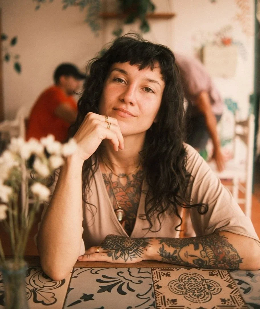 Eine Frau mit Tattoos sitzt in einem Café und blickt in die Kamera, während sie die Hand an ihr Kinn legt.