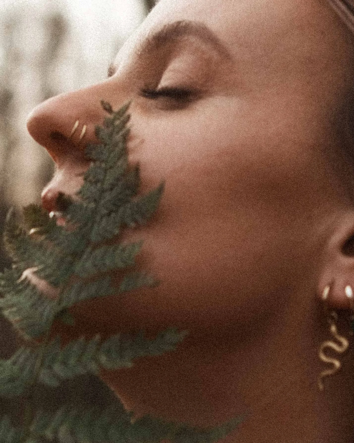 Nahaufnahme eines Frauengesichts mit geschlossenen Augen, eine grüne Pflanze bedeckt den unteren Teil des Gesichts, einen goldene Ohrring in der Ohrläppchen.