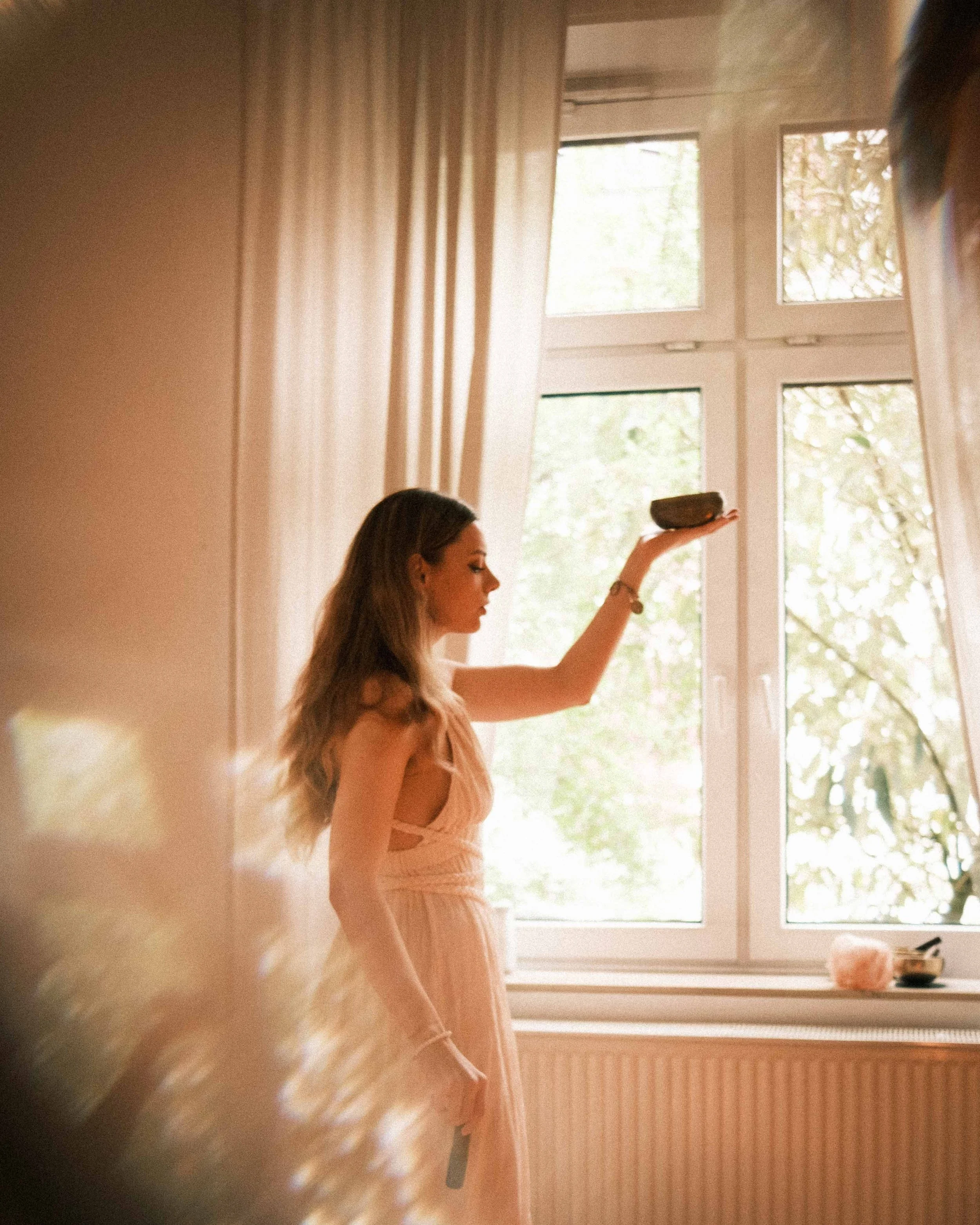 Frau in einem zeremoniellen Kleid hält eine Klangschale vor einem großen Fenster mit weißen Rahmen und transparenten Vorhängen.