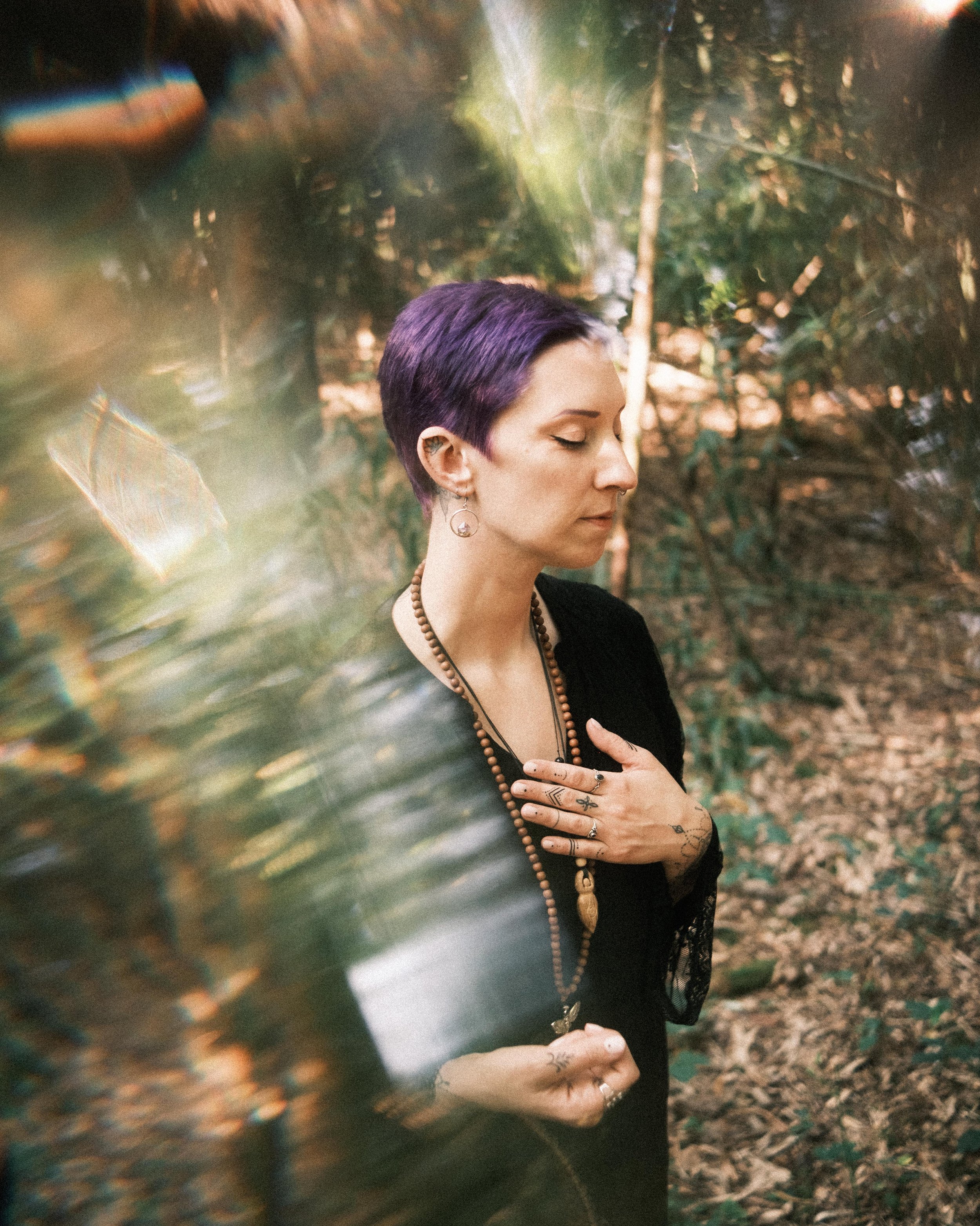 Eine Frau mit kurzen, violett gefärbten Haaren steht in einem Wald, ihre Augen sind geschlossen, sie berührt ihre Brust und trägt Schmuck und Tattoos.