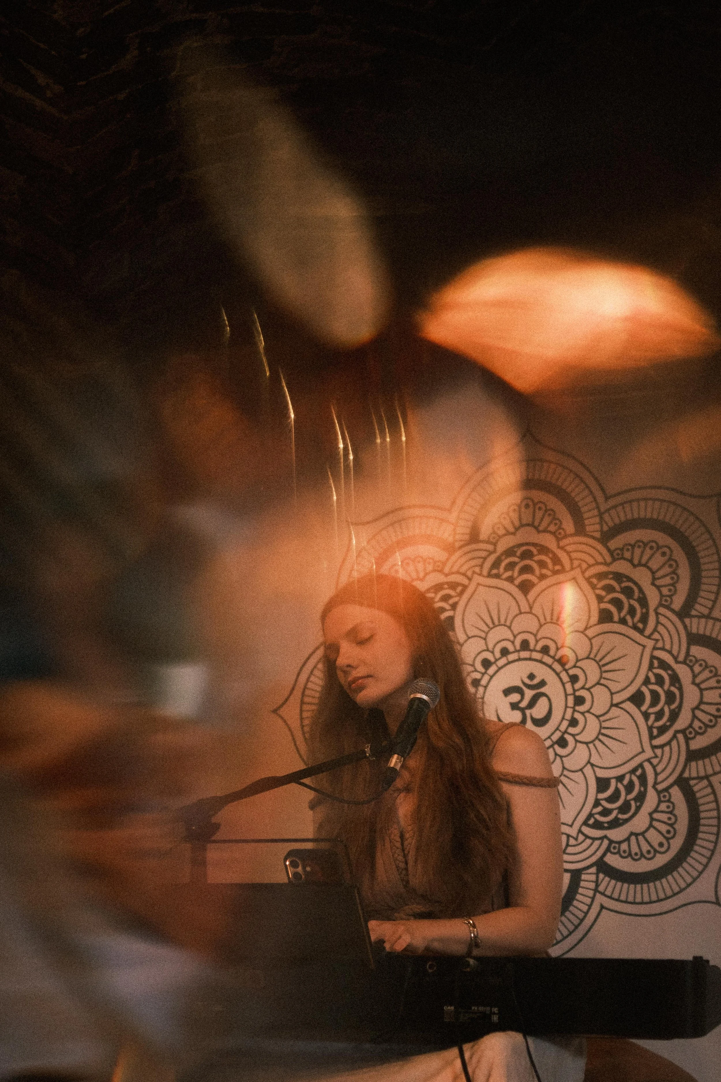 Eine junge Frau spielt Piano und singt in ein Mikrofon vor einem Mandala mit Om-Symbol im Hintergrund, in einer sanft und warm beleuchteten Umgebung.