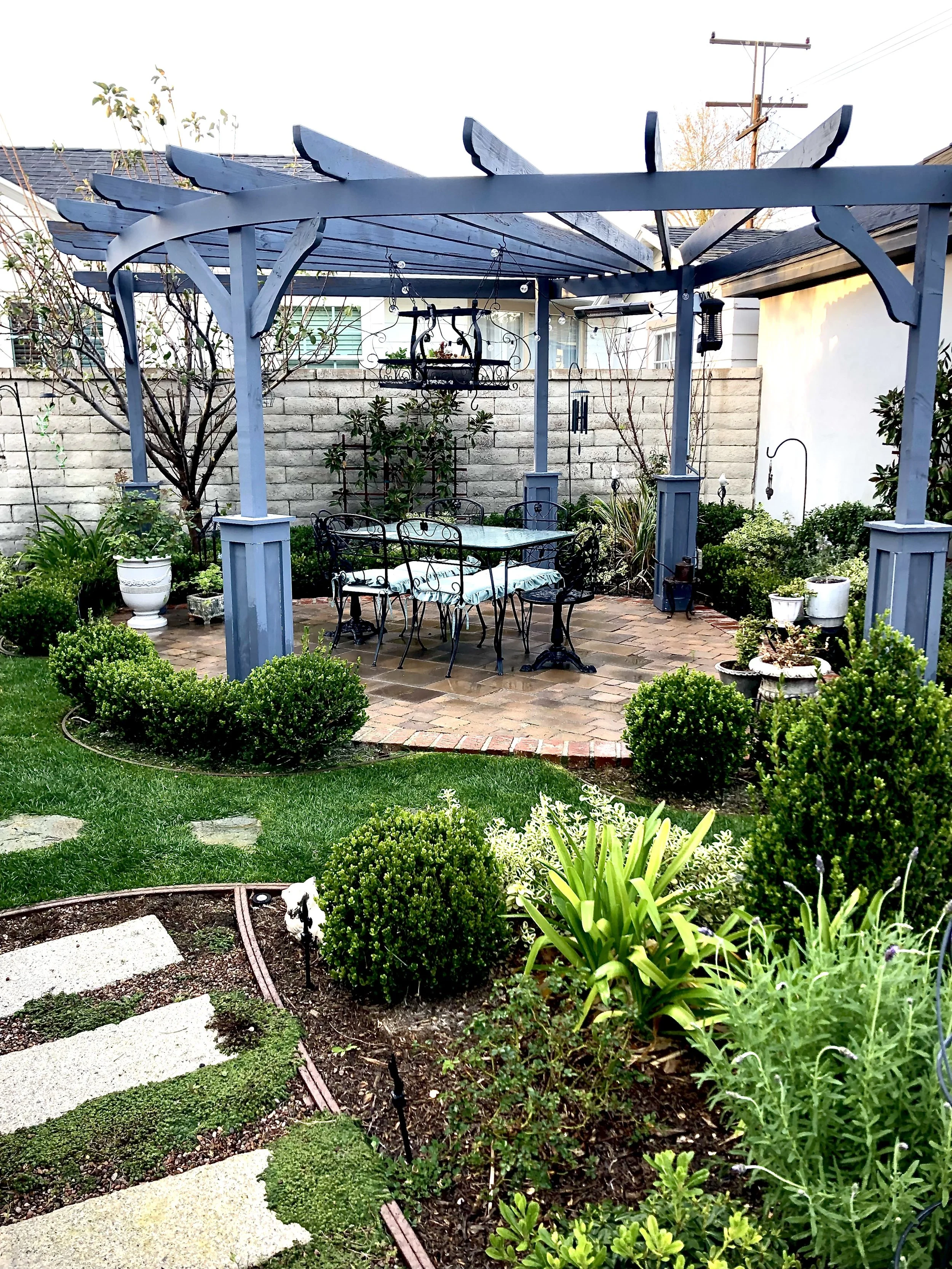 A painted pergola shelters a brick patio and dining area, surrounded by clipped boxwood and layered garden plantings