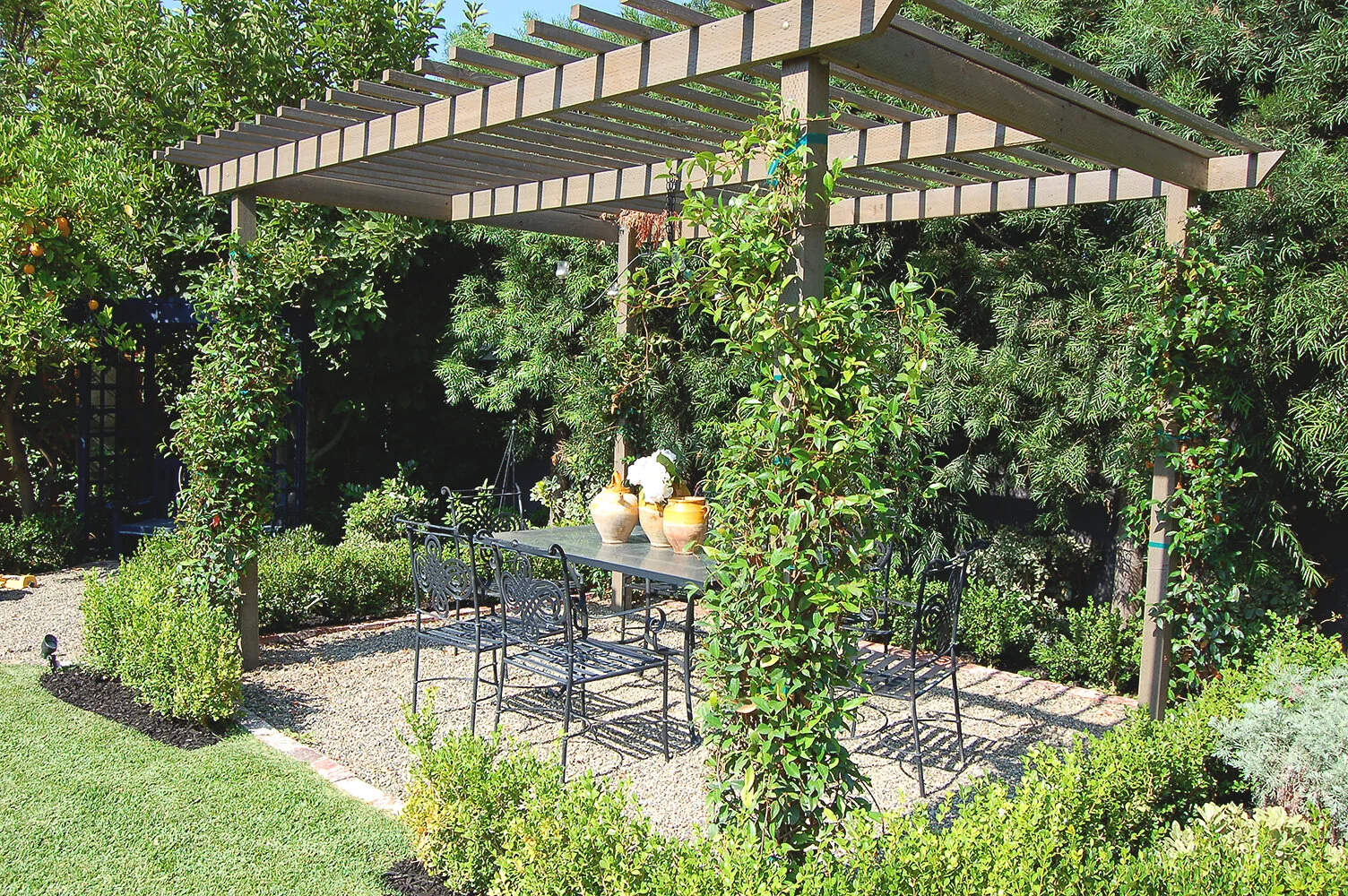 A shaded garden dining area beneath a vine-covered pergola creates an inviting outdoor room
