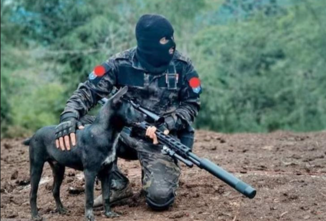 A person in military gear wearing a black ski mask, kneeling in dirt, holding a rifle, with a black dog standing in front of them, outdoors with green trees in the background.