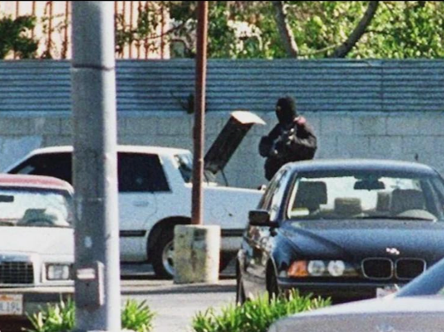 A person wearing a black hoodie and cap, standing behind a row of parked cars, with their arms crossed in a parking lot.