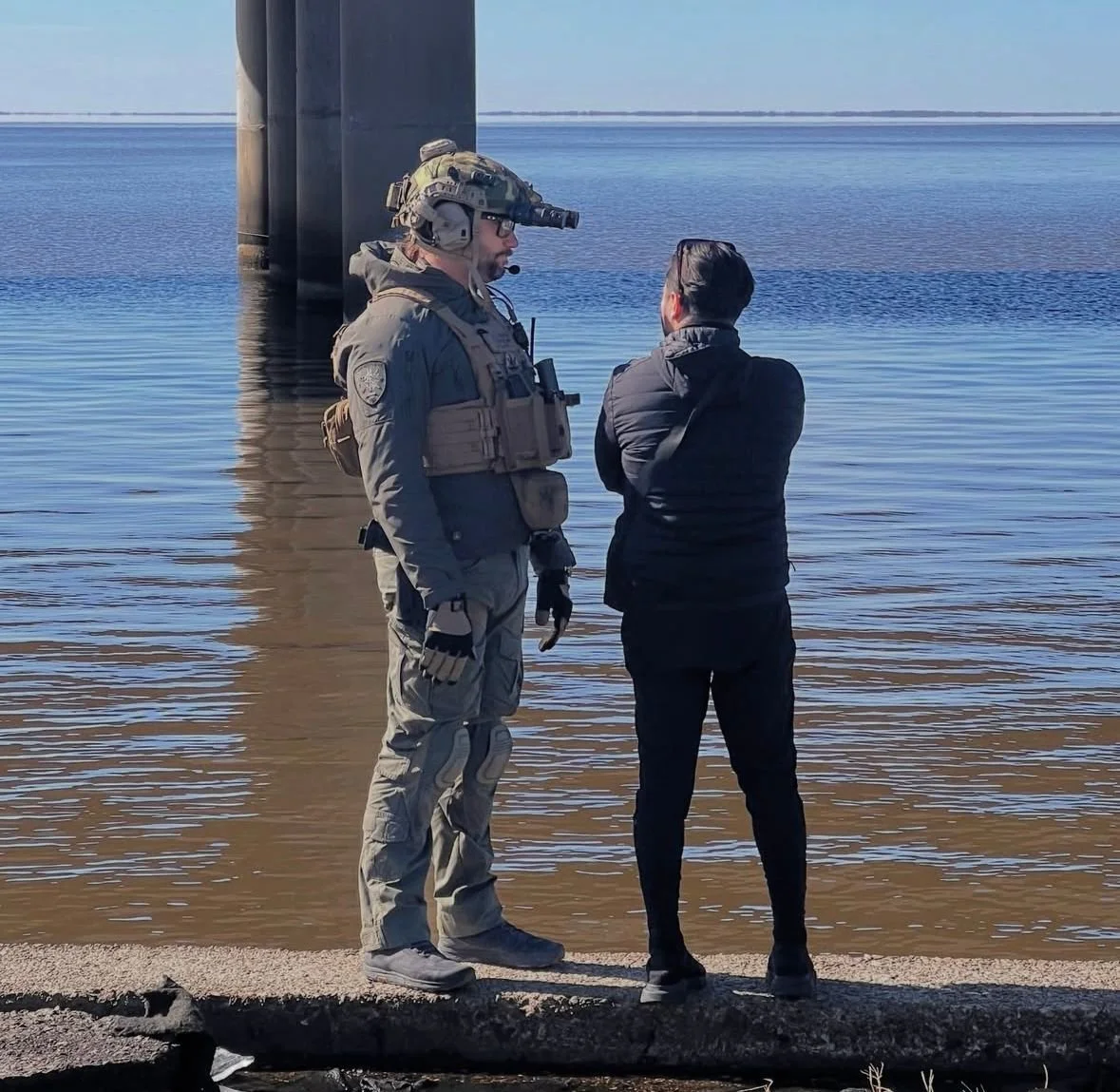 A man in military gear and a woman in black clothing stand on a pier by water, with a bridge in the background.
