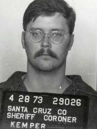 Black and white photo of a man with glasses and a mustache holding a mugshot board with his info.