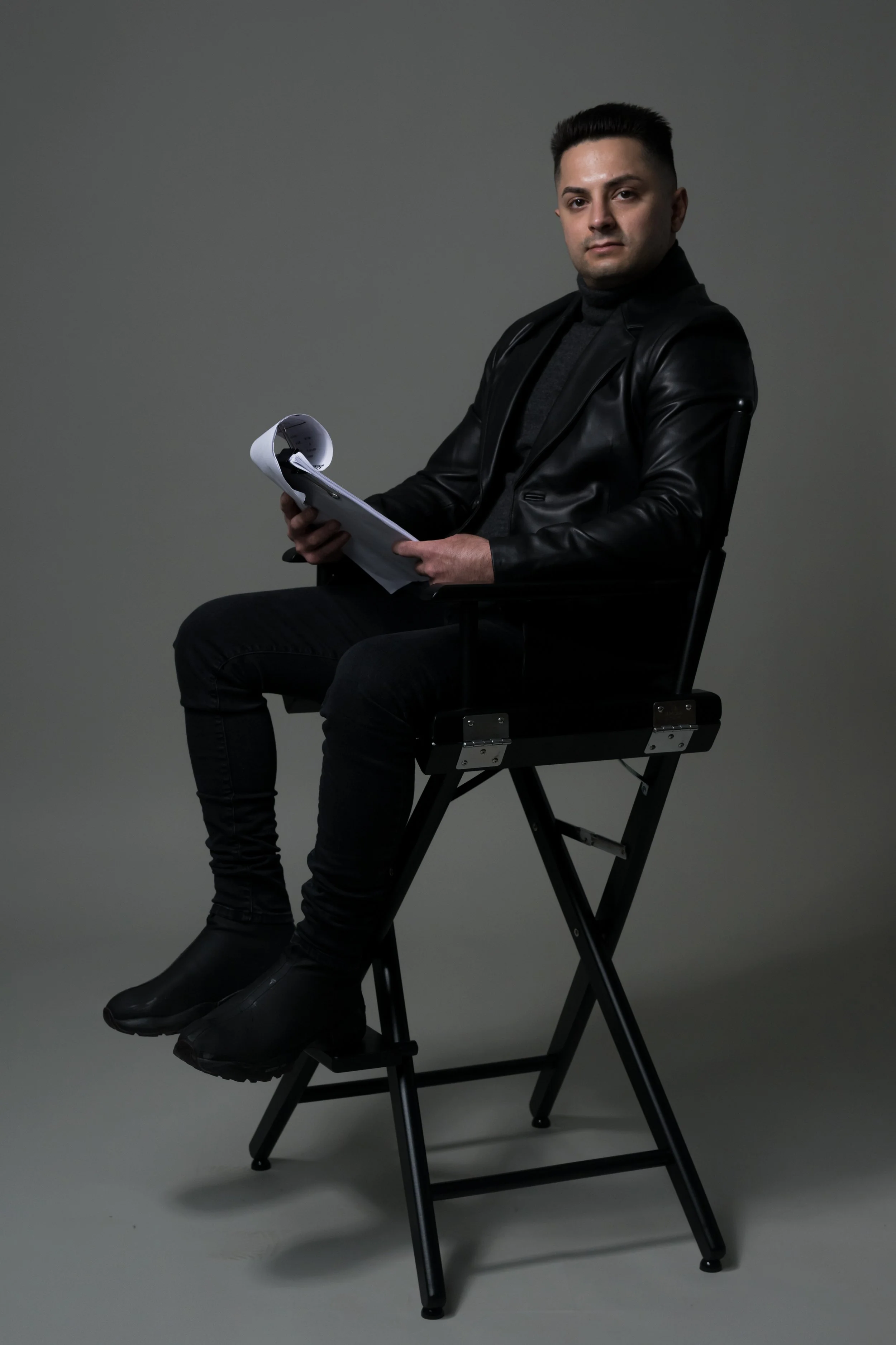 A man sitting on a high director's chair holding a script or paper, wearing a black leather jacket and dark pants against a plain background.