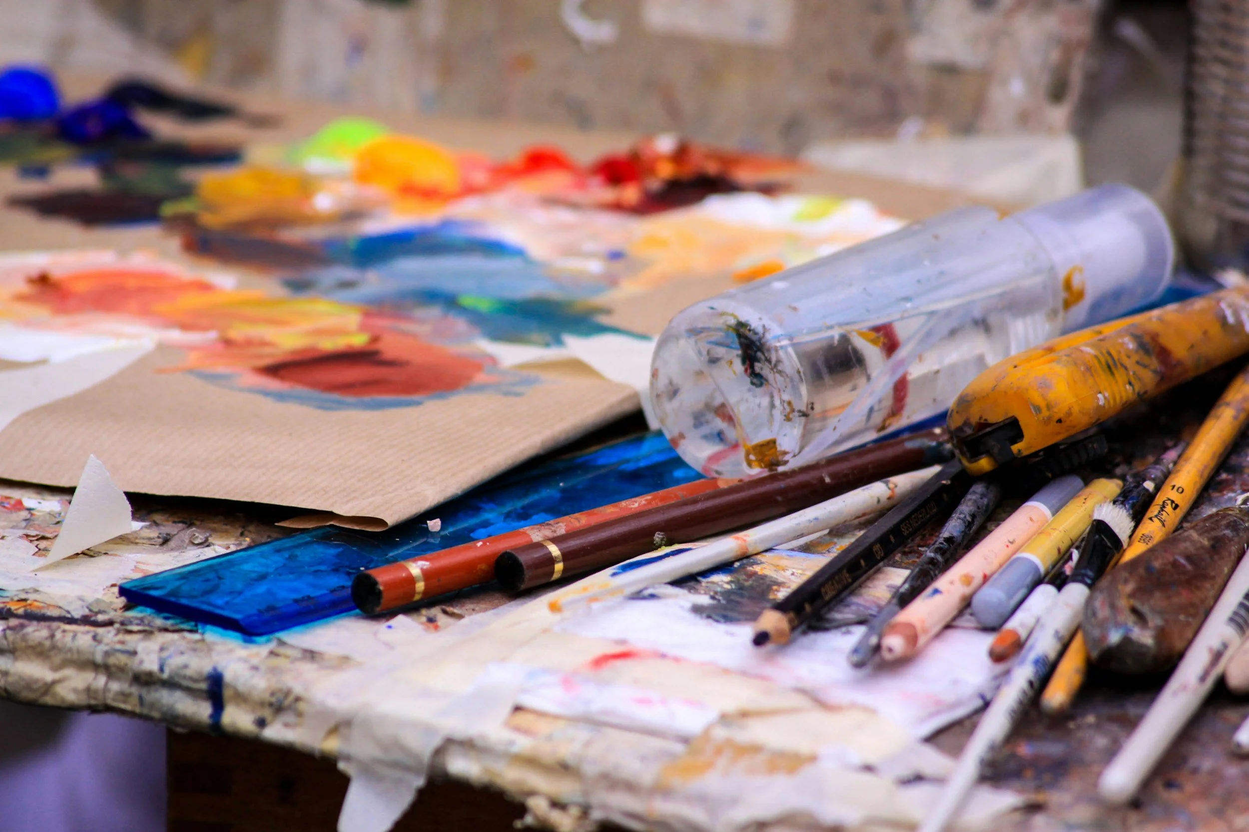 A cluttered art studio table with watercolor paints, brushes, a plastic container, and a palette, all covered in paint.