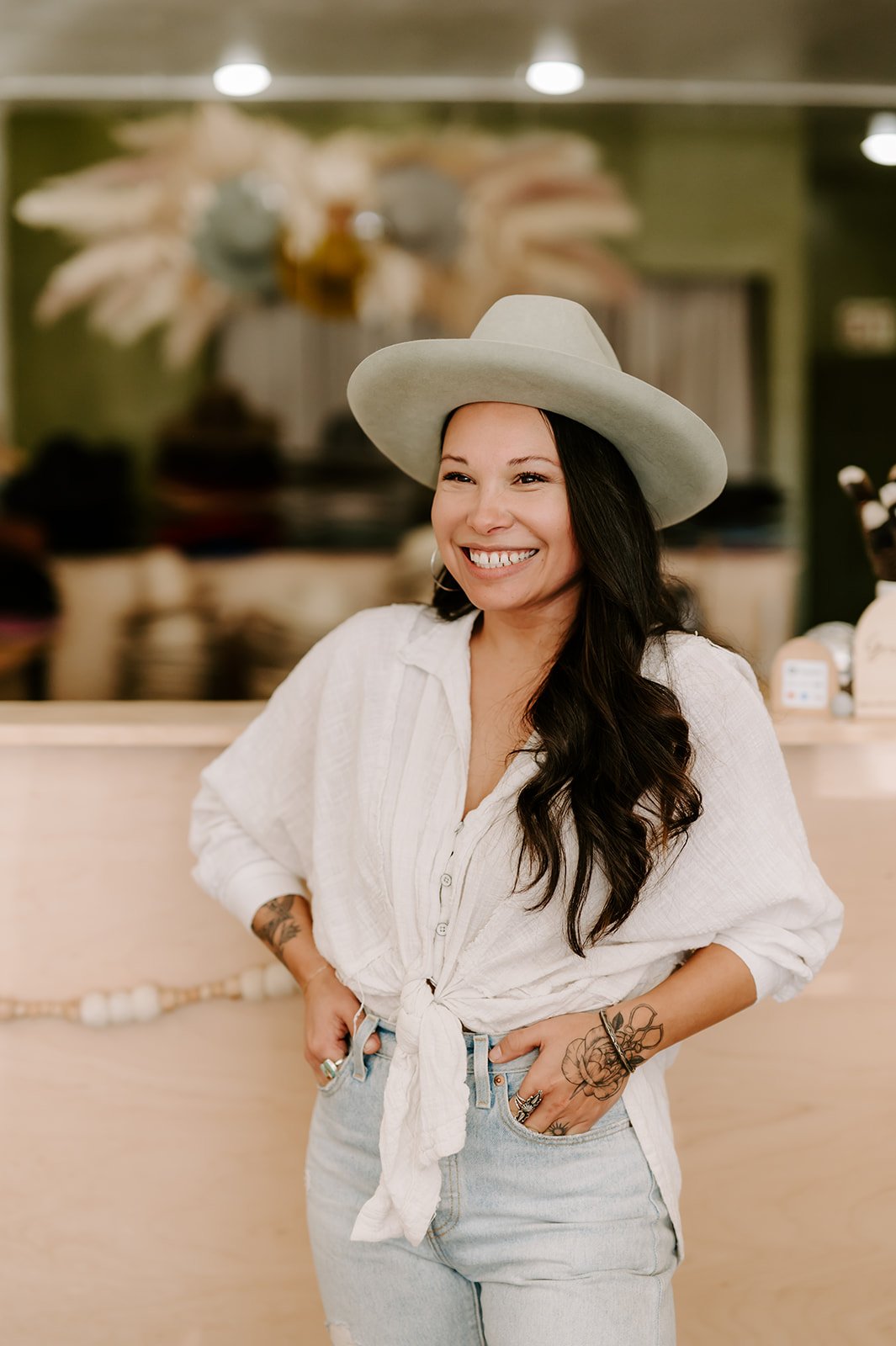 A smiling woman with long dark hair, wearing a white wide-brimmed hat, white button-up shirt tied at the waist, and light blue jeans, standing indoors with a blurred background.