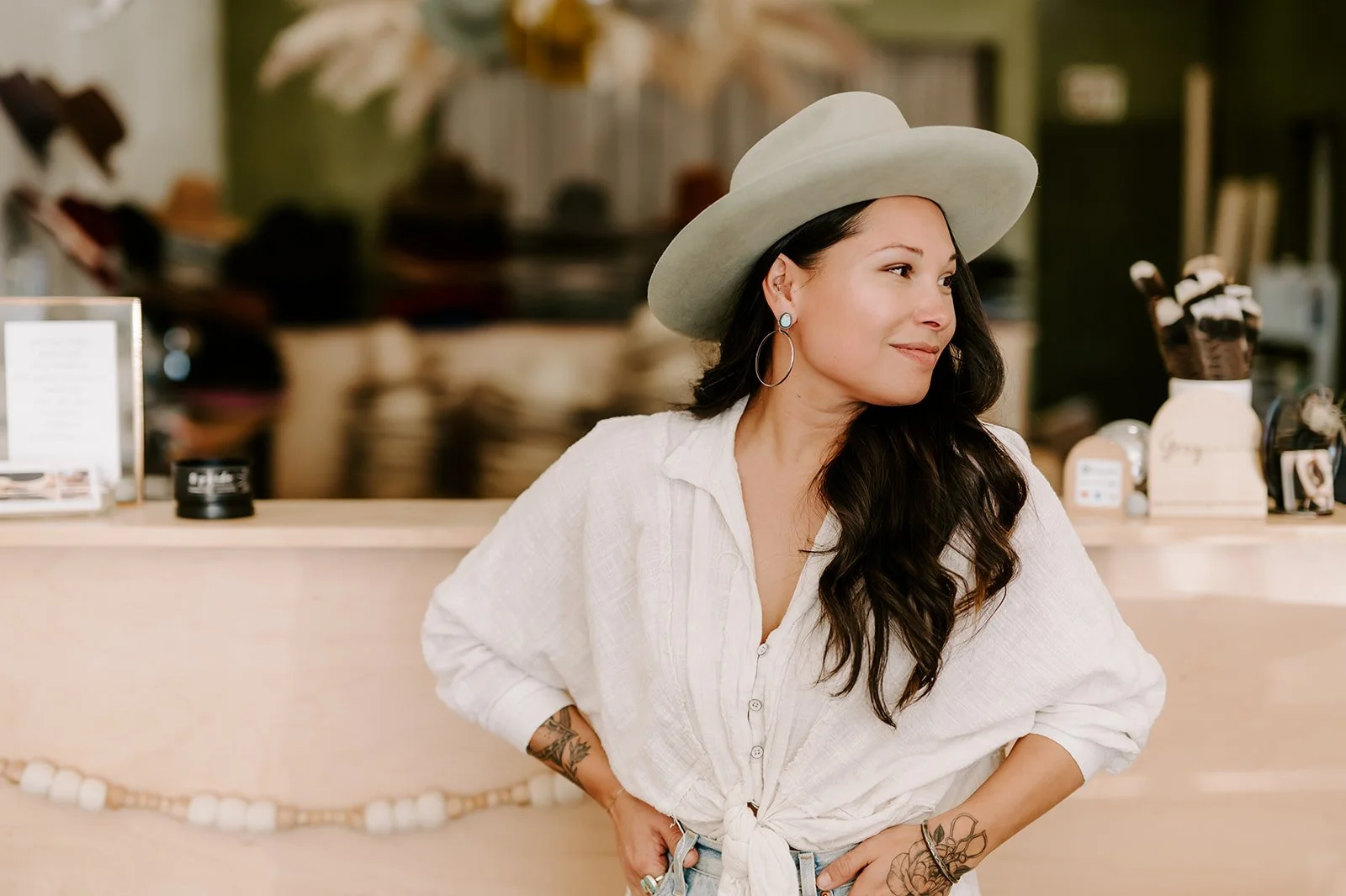 A woman with long dark hair wearing a light-colored wide-brimmed hat, hoop earrings, and a white tied-up blouse, standing at a counter in a hat shop.