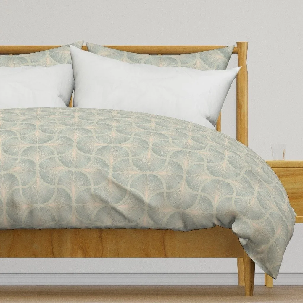A bed with a light wood frame, white pillows, and a decorative duvet with a light blue and beige pattern. Designed by Wild Geranium Design.