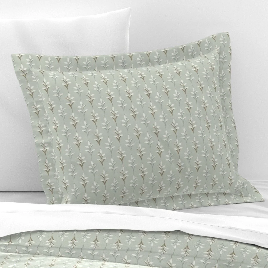 A decorative bed pillow with a light-colored pattern of small, repeated leaf designs, placed on a white bed with a white pillow behind it. Designed by Wild Geranium Design.